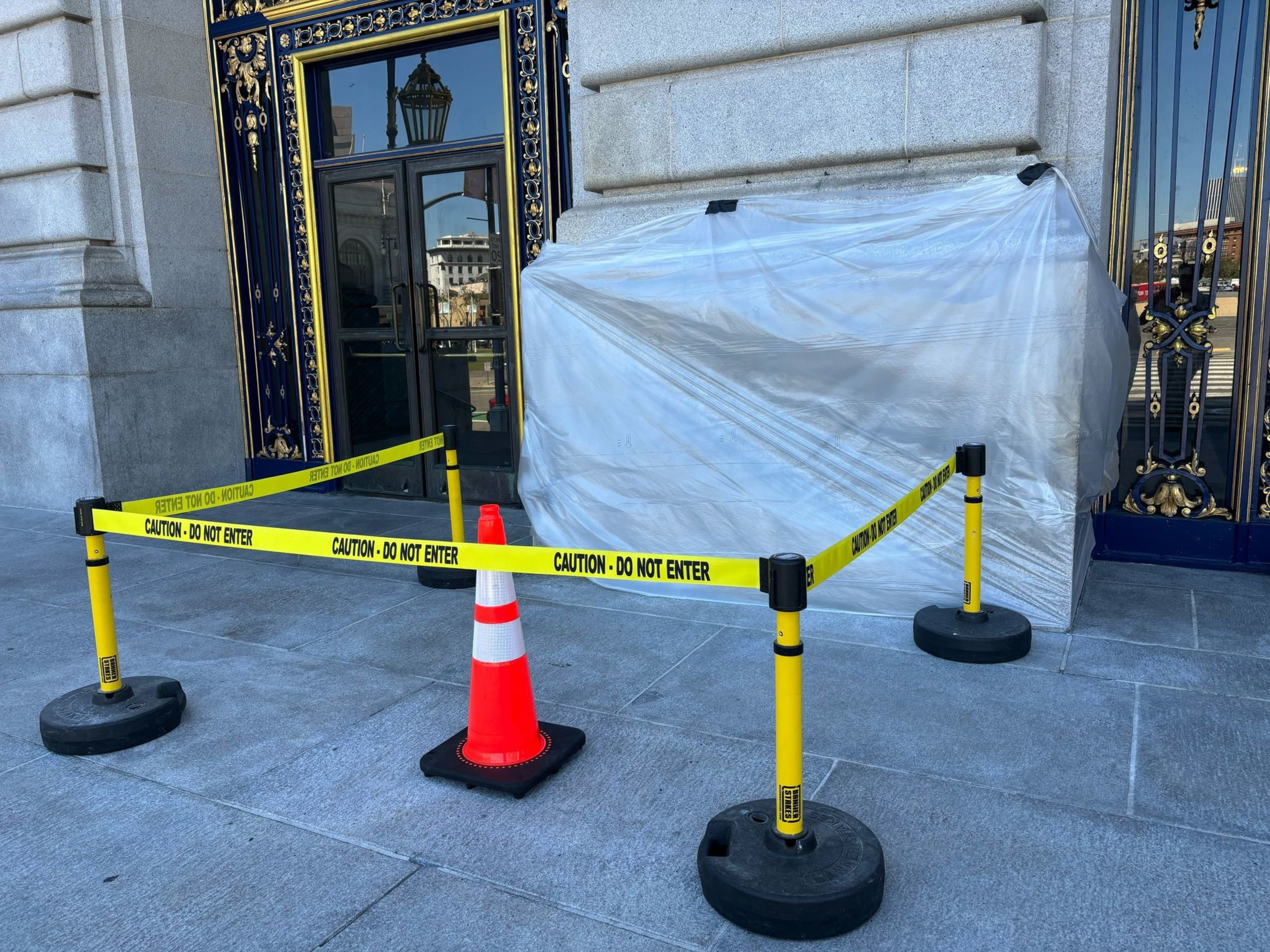 A doorway is blocked by "Caution - Do Not Enter" tape and a traffic cone. A large object covered in plastic is situated next to the door.