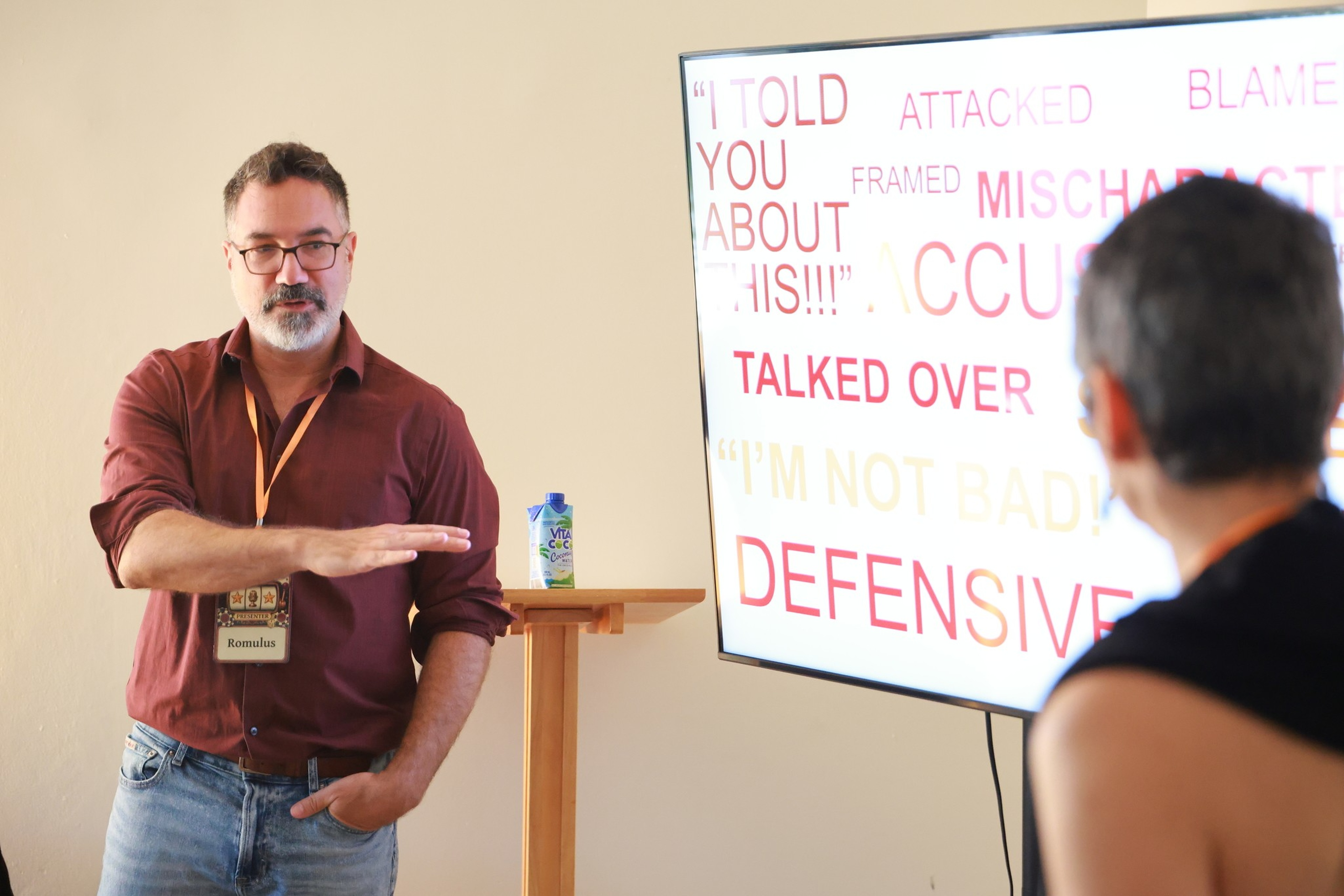 A man wearing glasses and a red shirt gestures while speaking to a woman, with a screen showing words like “DEFENSIVE” and “I TOLD YOU ABOUT THIS!!!” behind him.