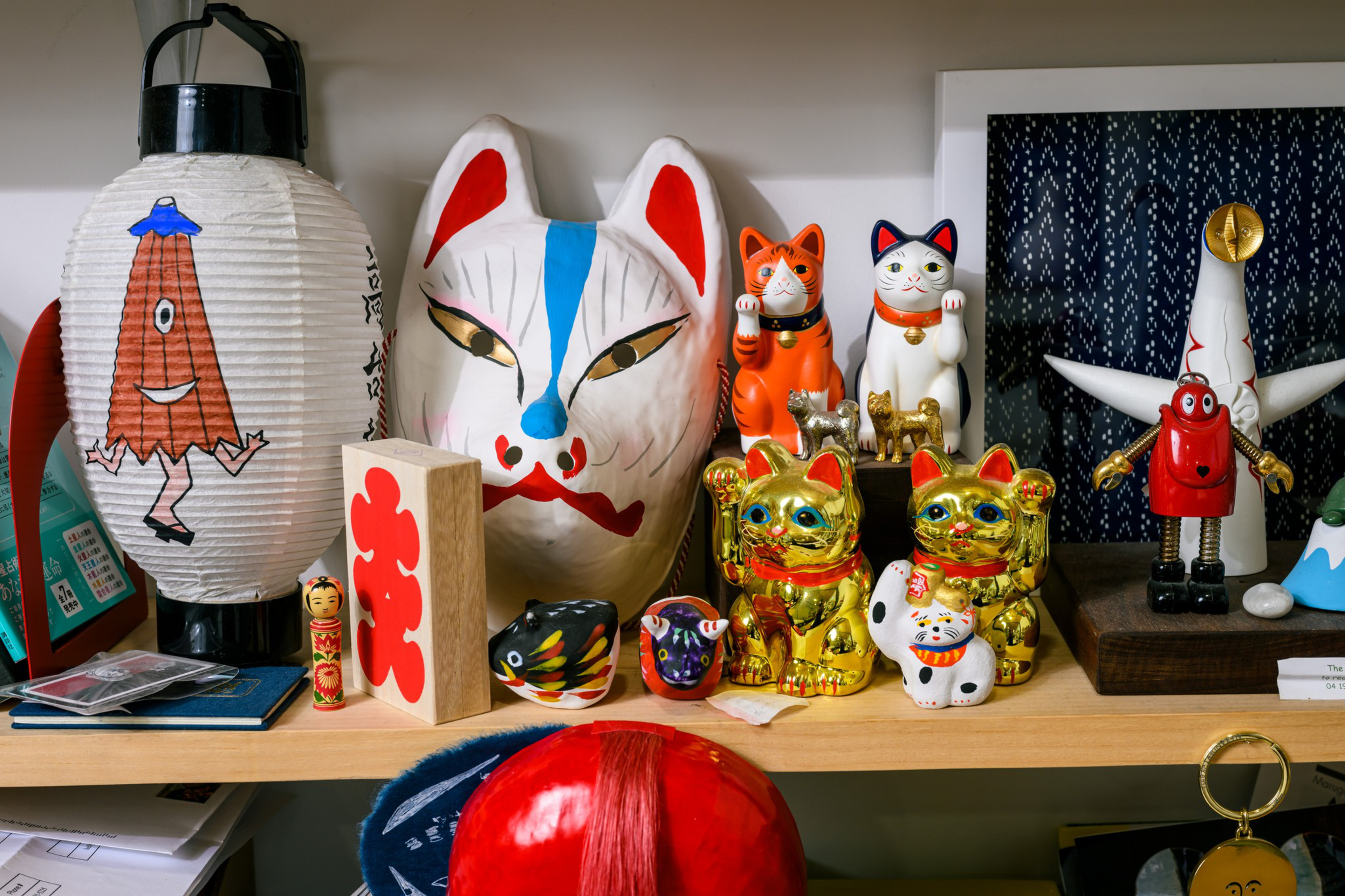 The image shows a shelf with various Japanese items, including maneki-neko cats, a fox mask, a paper lantern, and a small toy robot.