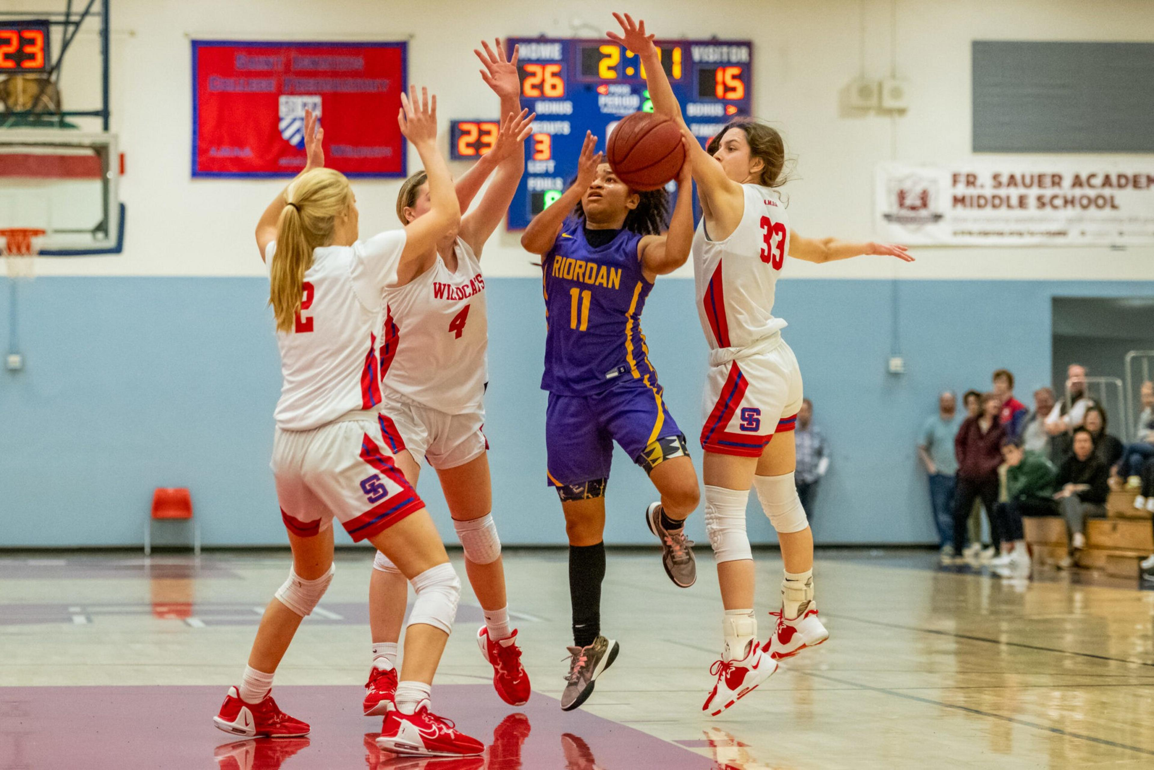 A basketball player in a purple and yellow uniform attempts a shot while three defenders in white and red jump to block her.