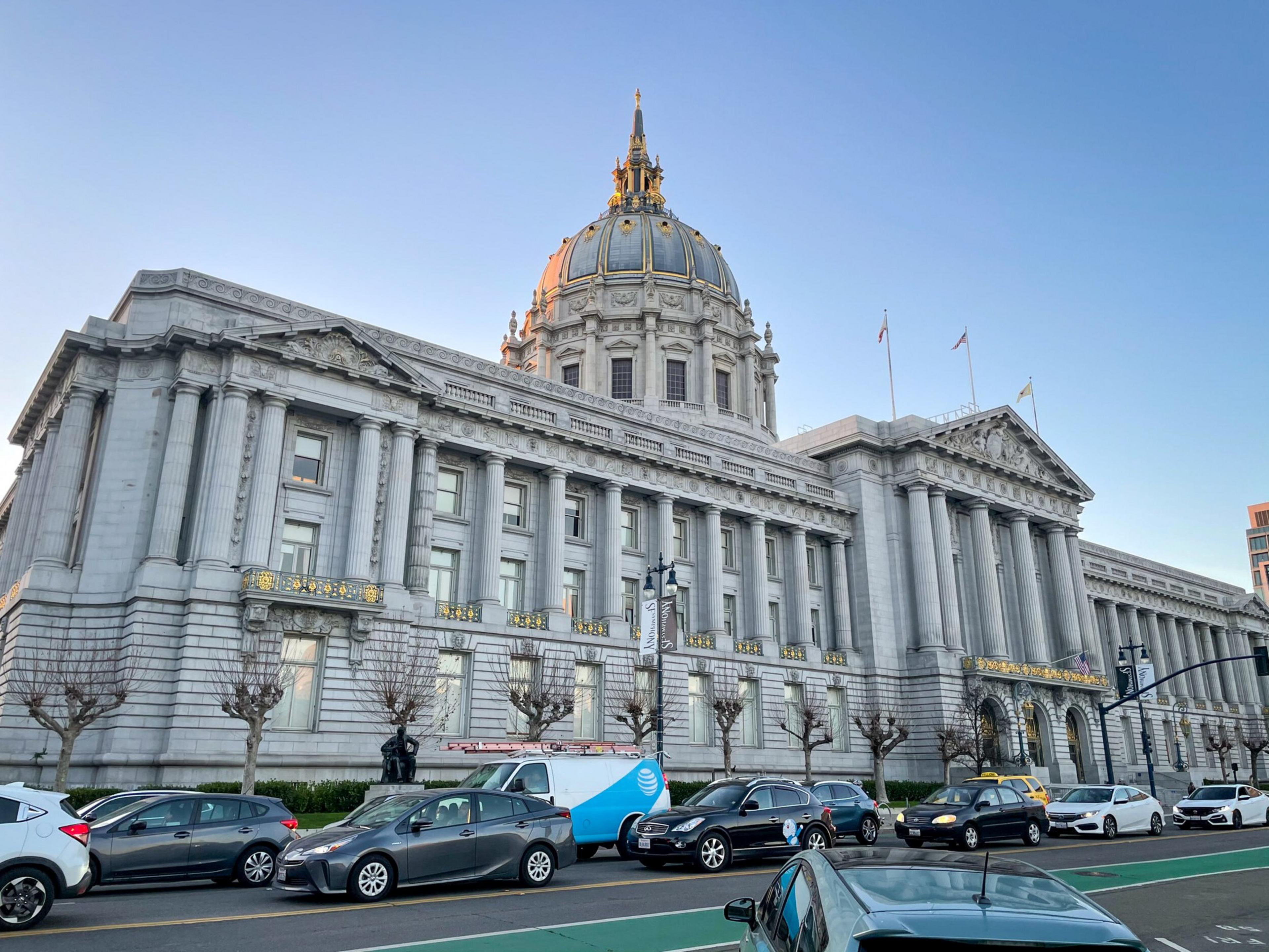 A large neoclassical building with a tall dome and columns stands behind a busy street filled with parked cars and leafless trees under a clear blue sky.