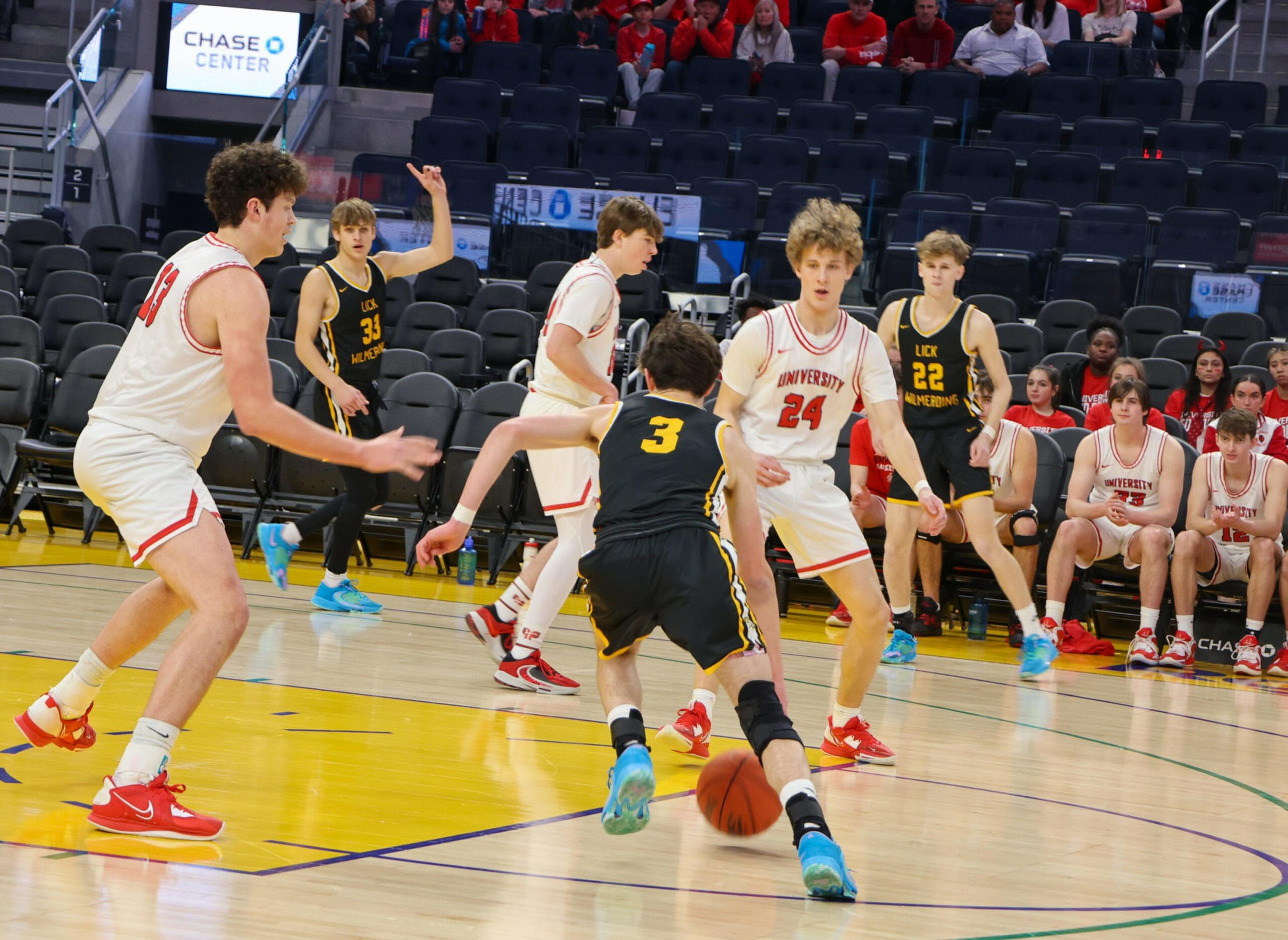A basketball player in a black jersey dribbles the ball while four players in white jerseys defend him on the court near the sidelines.