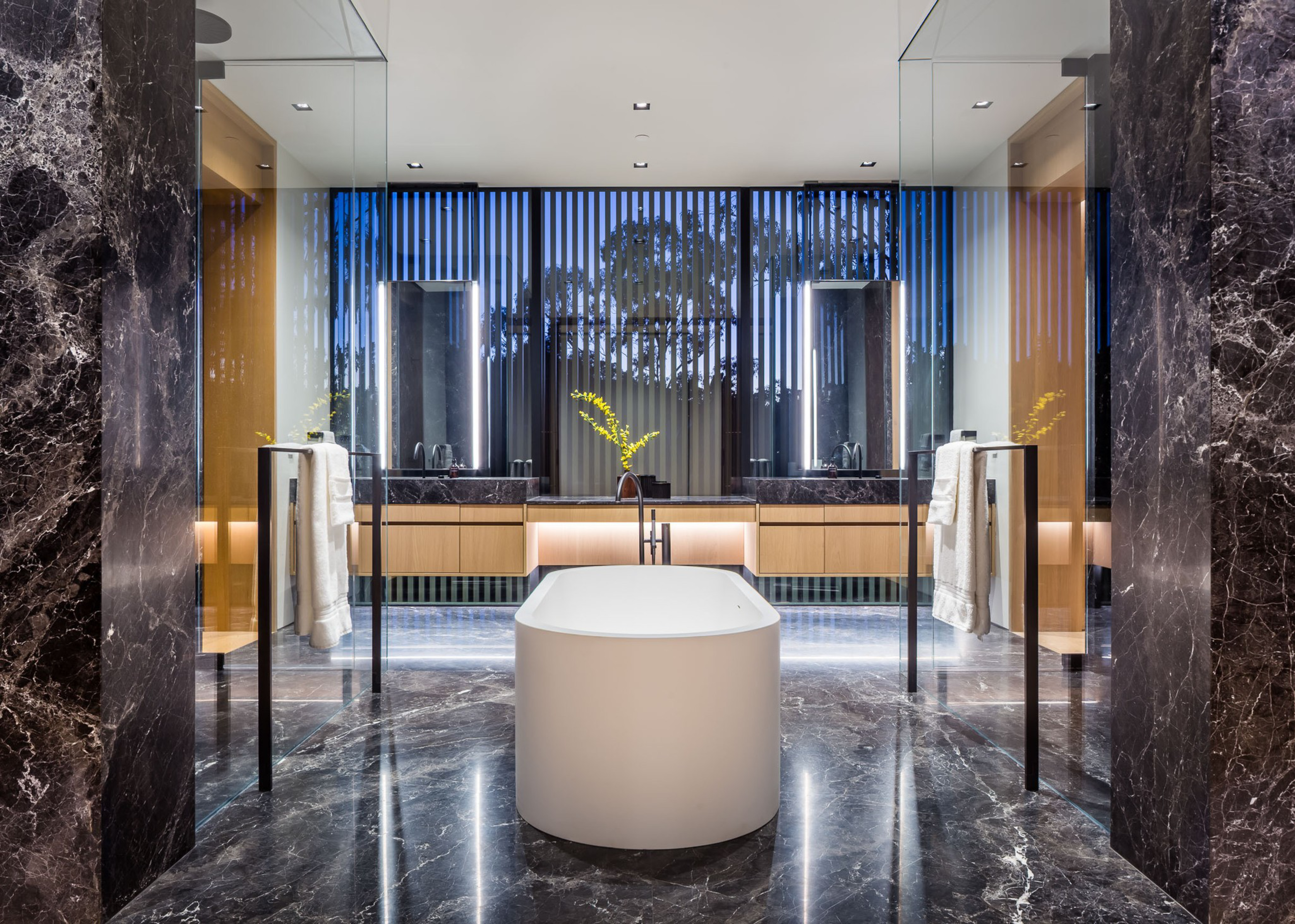 A modern bathroom features a central white freestanding bathtub on shiny dark marble flooring, with matching marble walls, twin sinks, and vertical blinds.