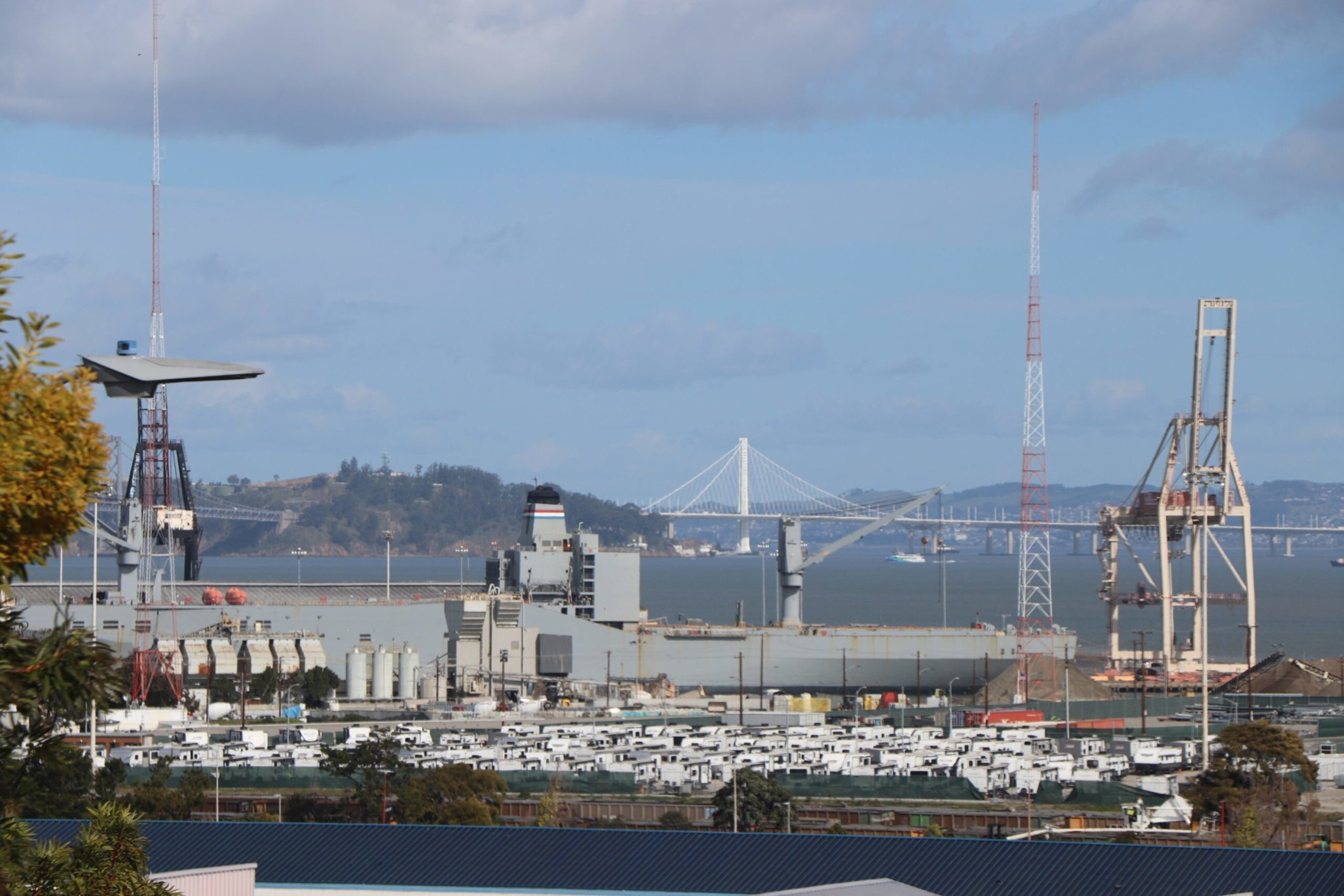 A large port facility with cranes and a ship docked, rows of white vehicles in the foreground, a mountainous island and a suspension bridge in the background.