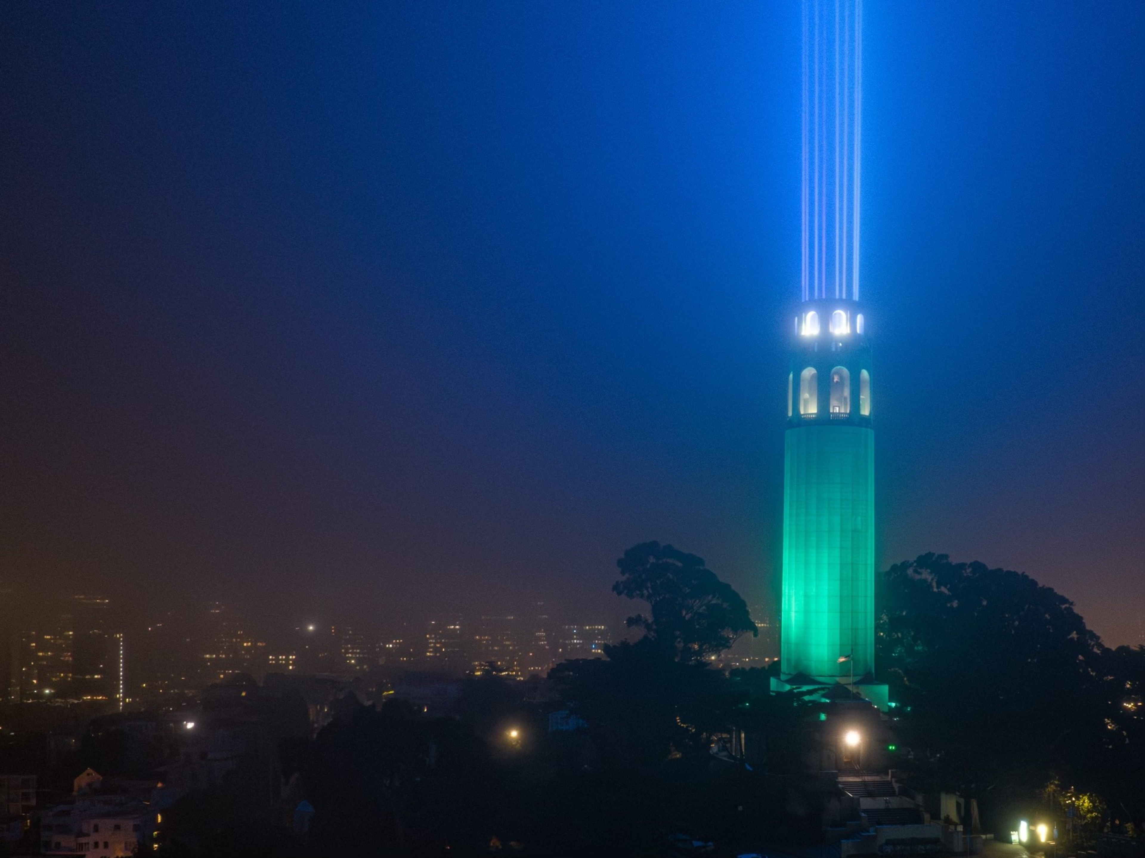 A tall tower illuminated with green light beams shooting upwards at night, surrounded by dark trees and city lights in the background.