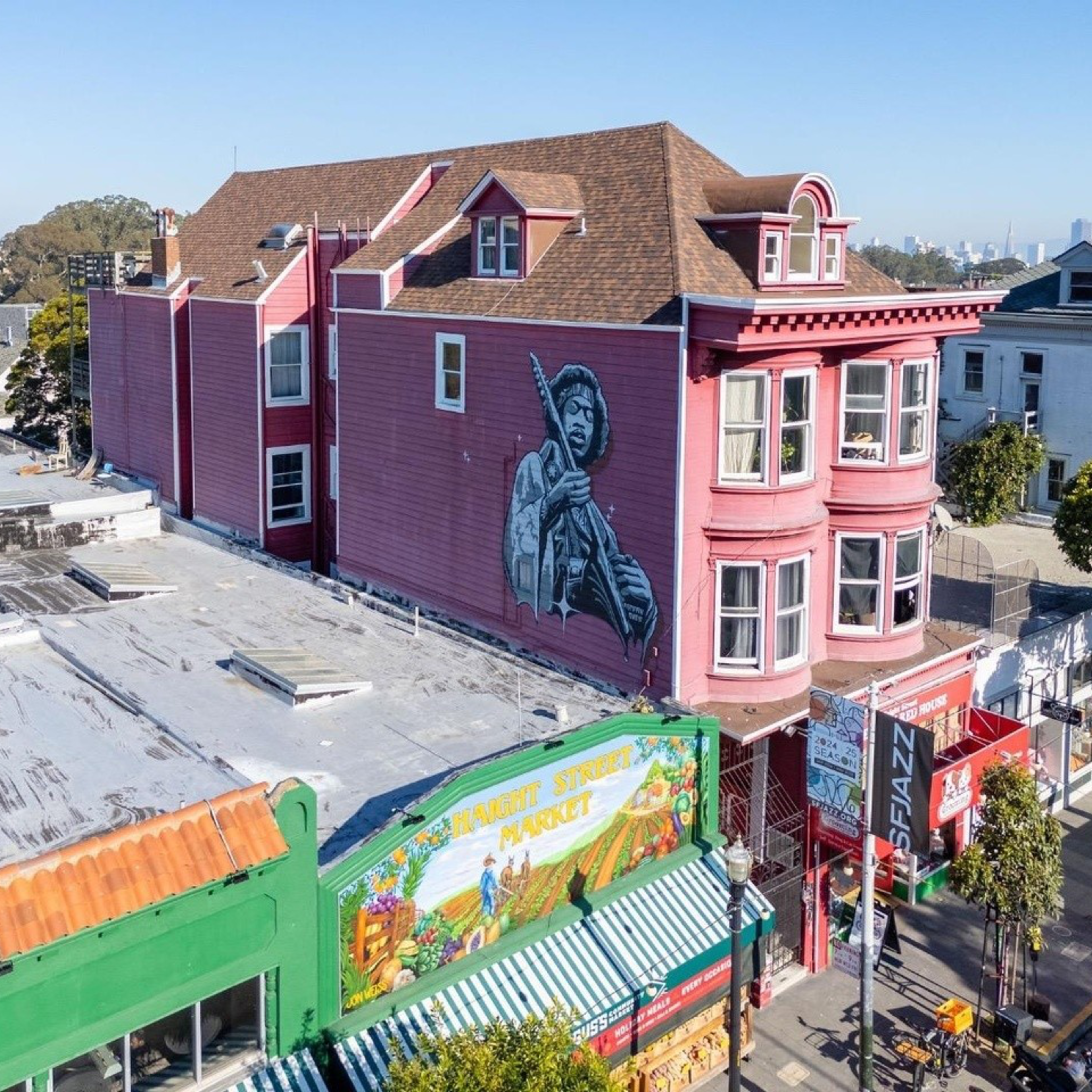 A mural of a musician holding a guitar is painted on a red building. Below, there's a colorful market with a bright striped awning and a few storefronts.