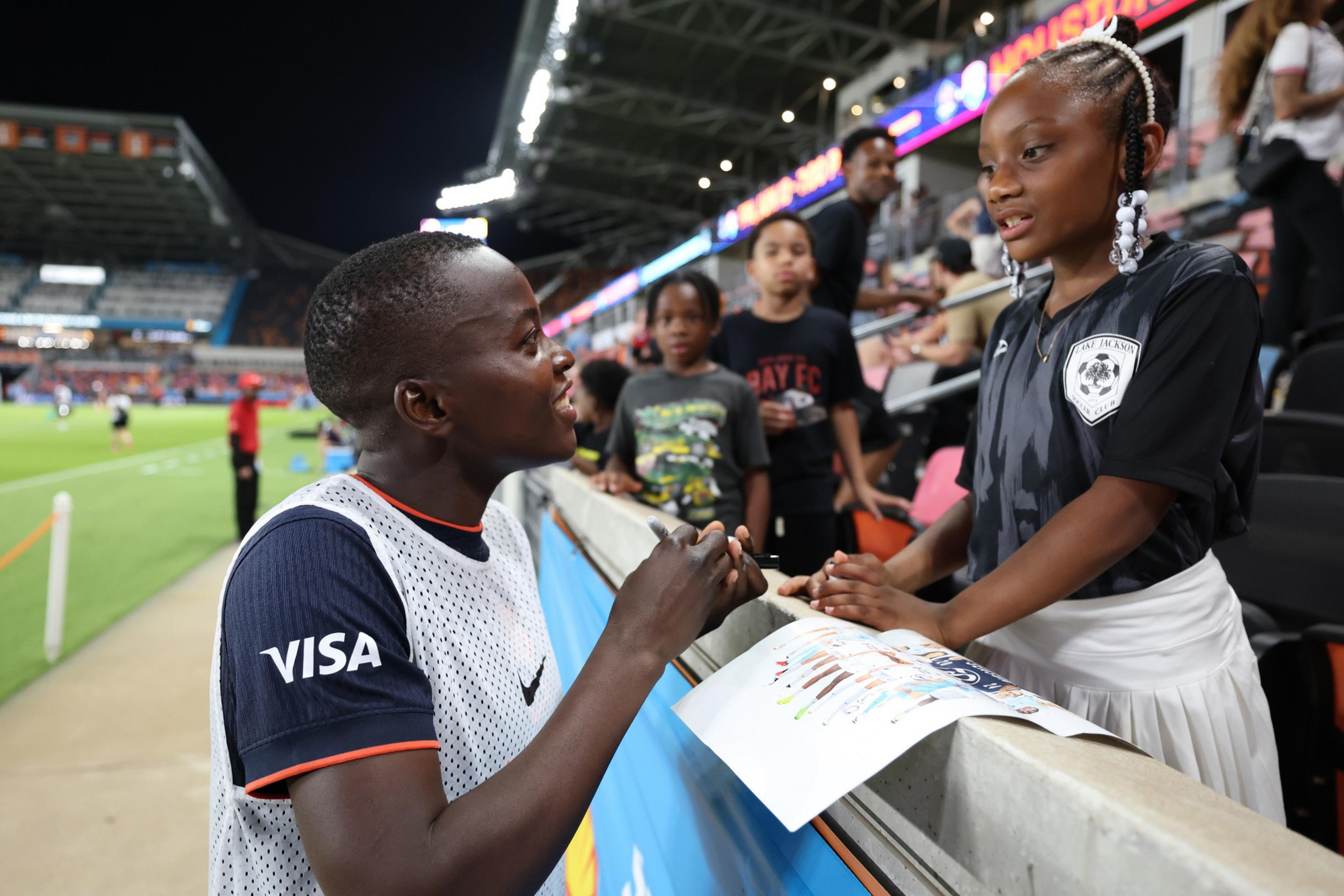 A young soccer player in a white vest talks and holds hands with a girl in a black jersey while fans watch from stadium seats nearby.