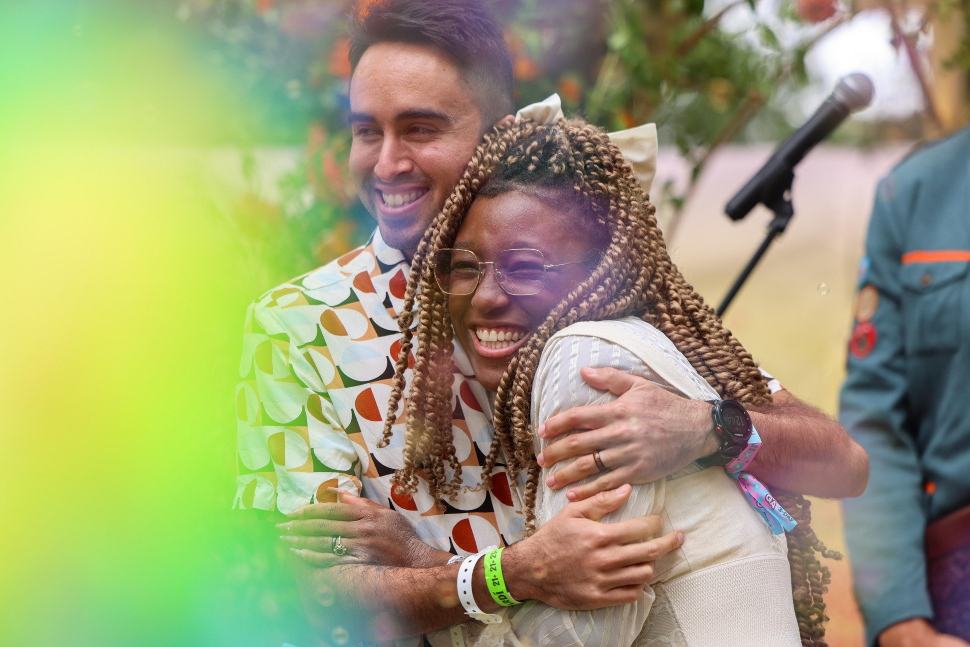 A man and woman are hugging joyfully, surrounded by colorful, blurred foliage. The woman has braided hair and glasses, while the man wears a patterned shirt.
