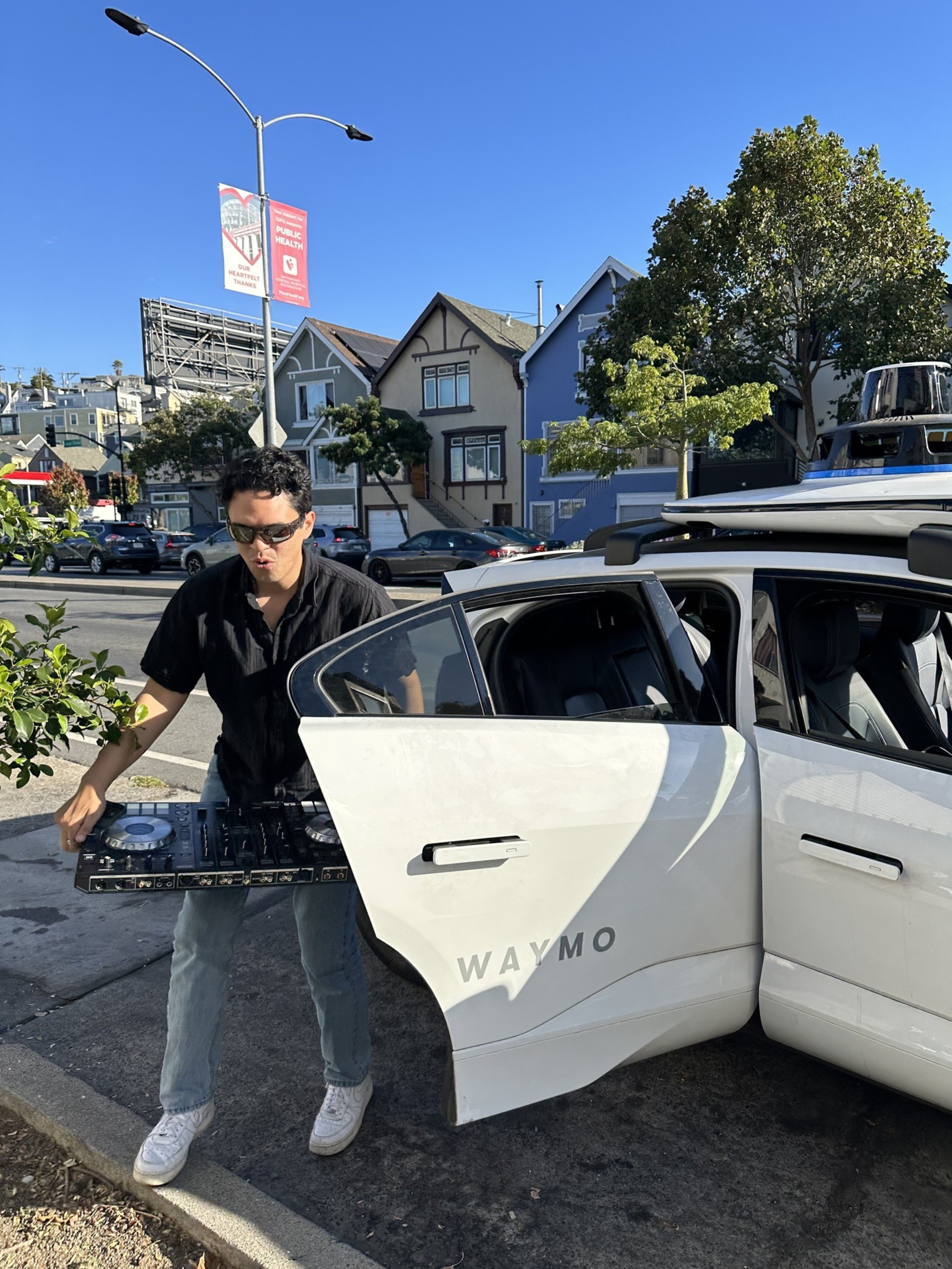 A person carrying DJ equipment steps out of a white car labeled &quot;Waymo&quot; on a sunny street with colorful houses and a clear blue sky in the background.