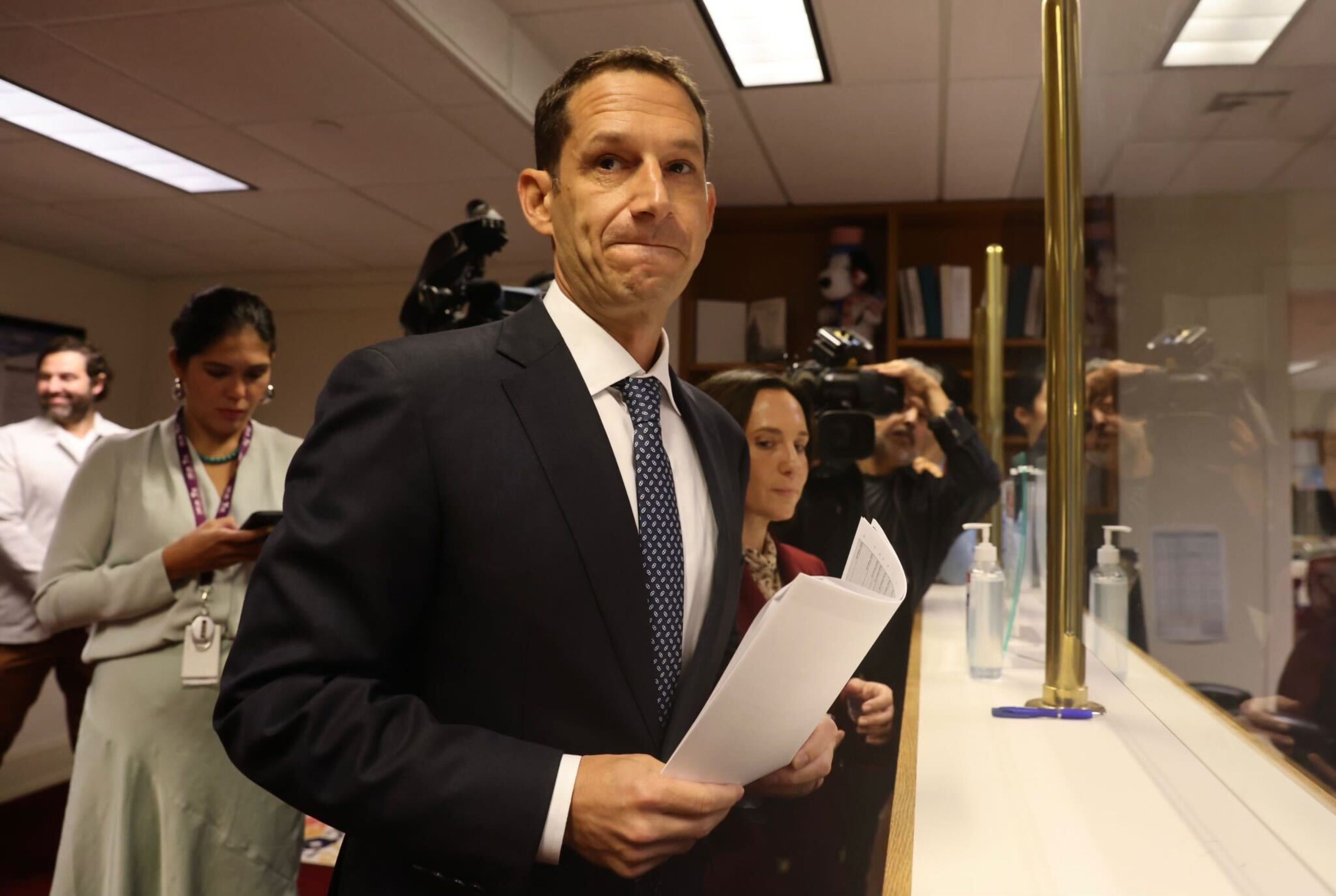 A man in a suit holds papers at a counter, surrounded by media personnel with cameras. A woman behind him looks at her phone. Bottles of sanitizer are on the counter.