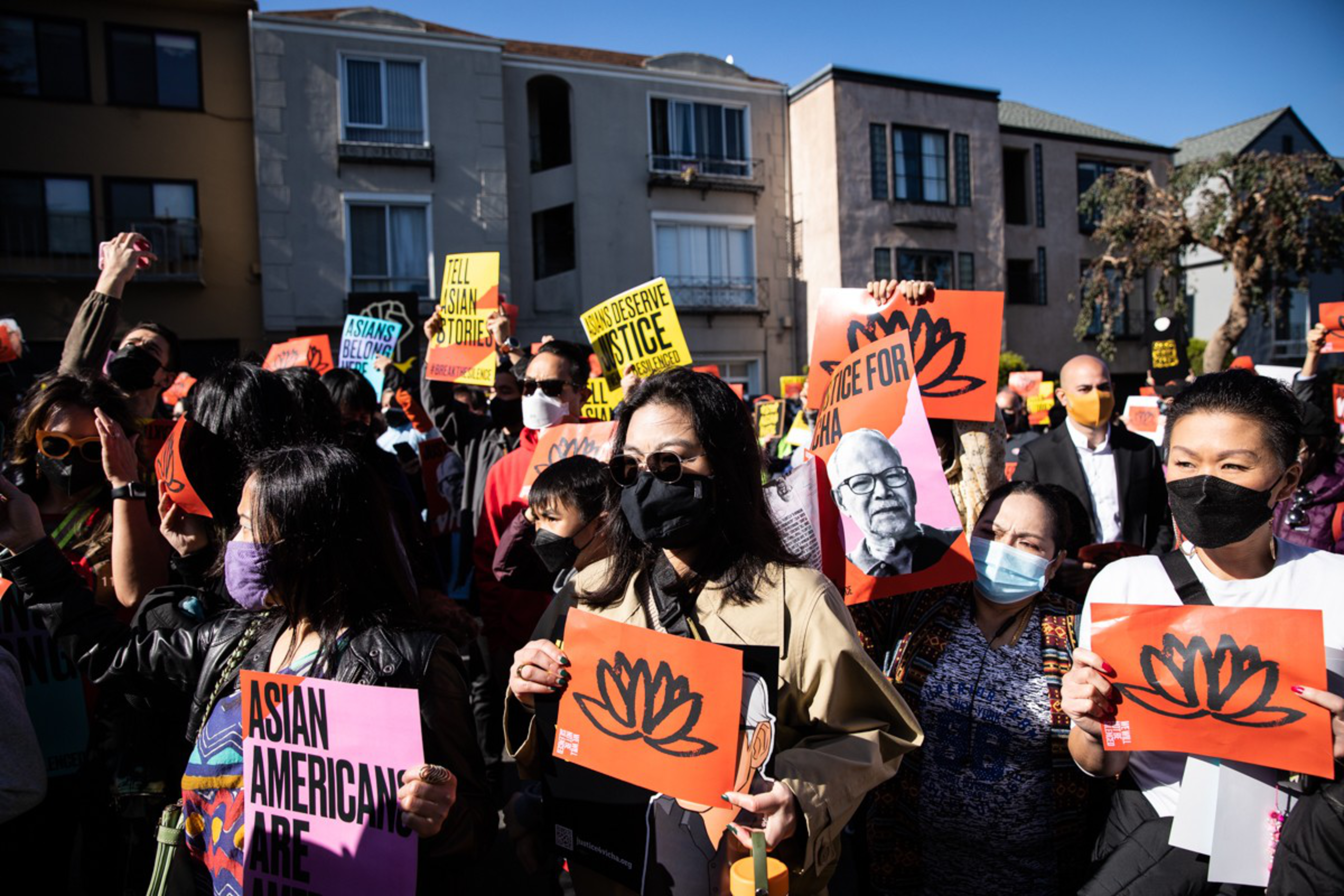 A diverse group of people wearing masks hold signs advocating for Asian American justice and rights in a sunny urban protest setting.