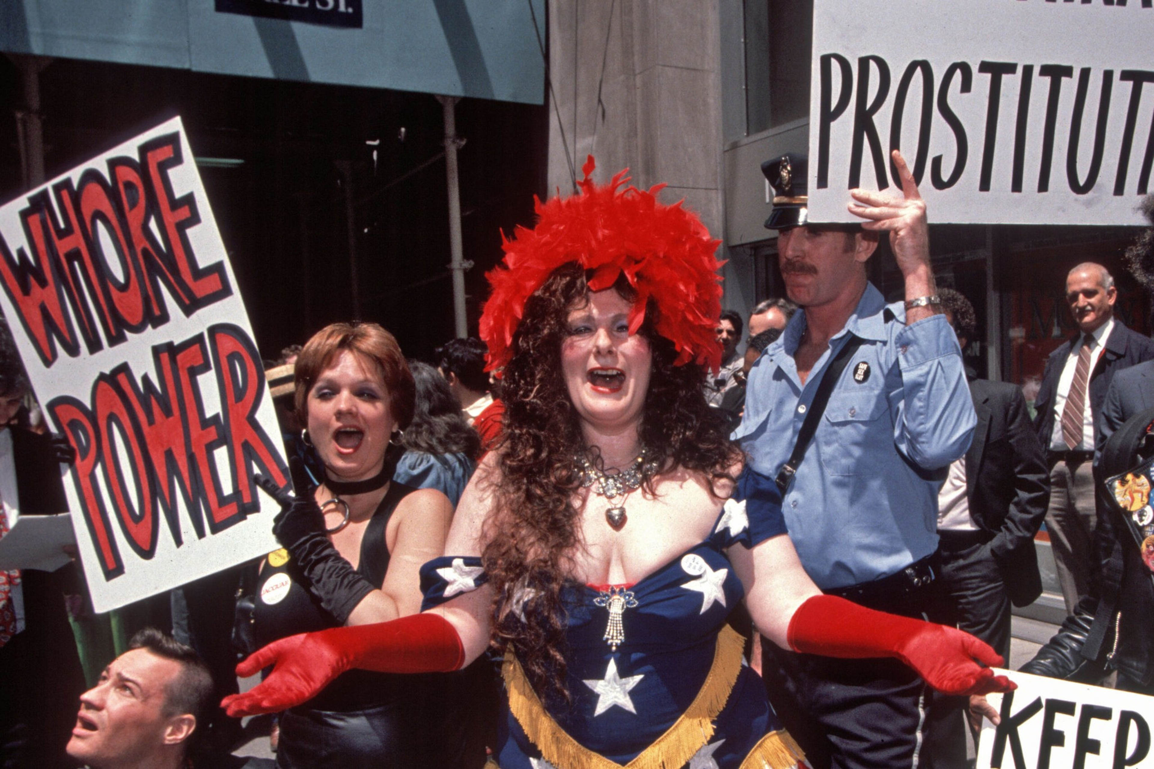 A woman in a star-spangled dress and red feathered hat gestures widely while others hold signs reading "WHORE POWER" and "PROSTITUT". A police officer stands nearby.