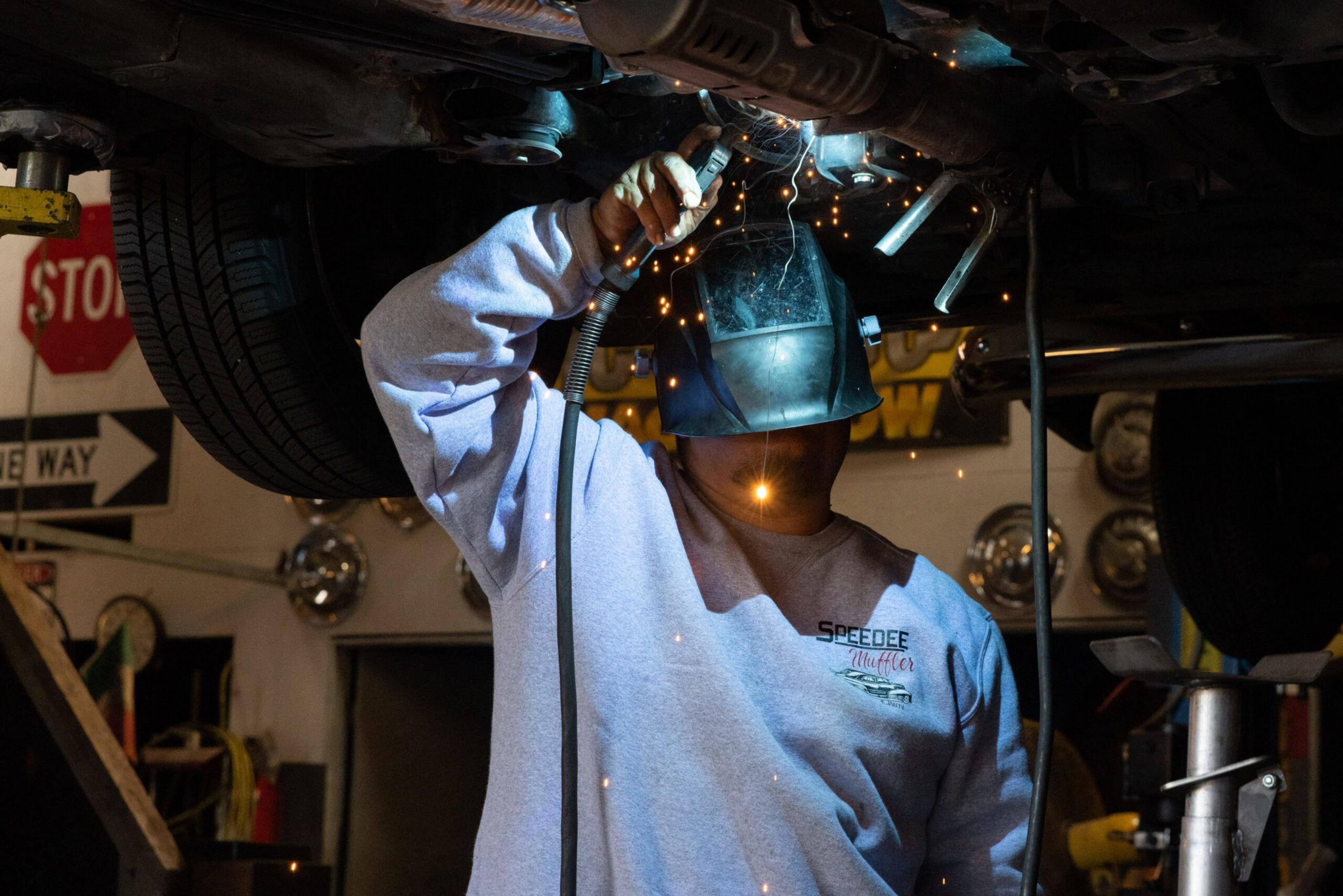 A person wearing a welding helmet and sweatshirt sparks while welding under a raised vehicle in a garage with signs and car parts in the background.