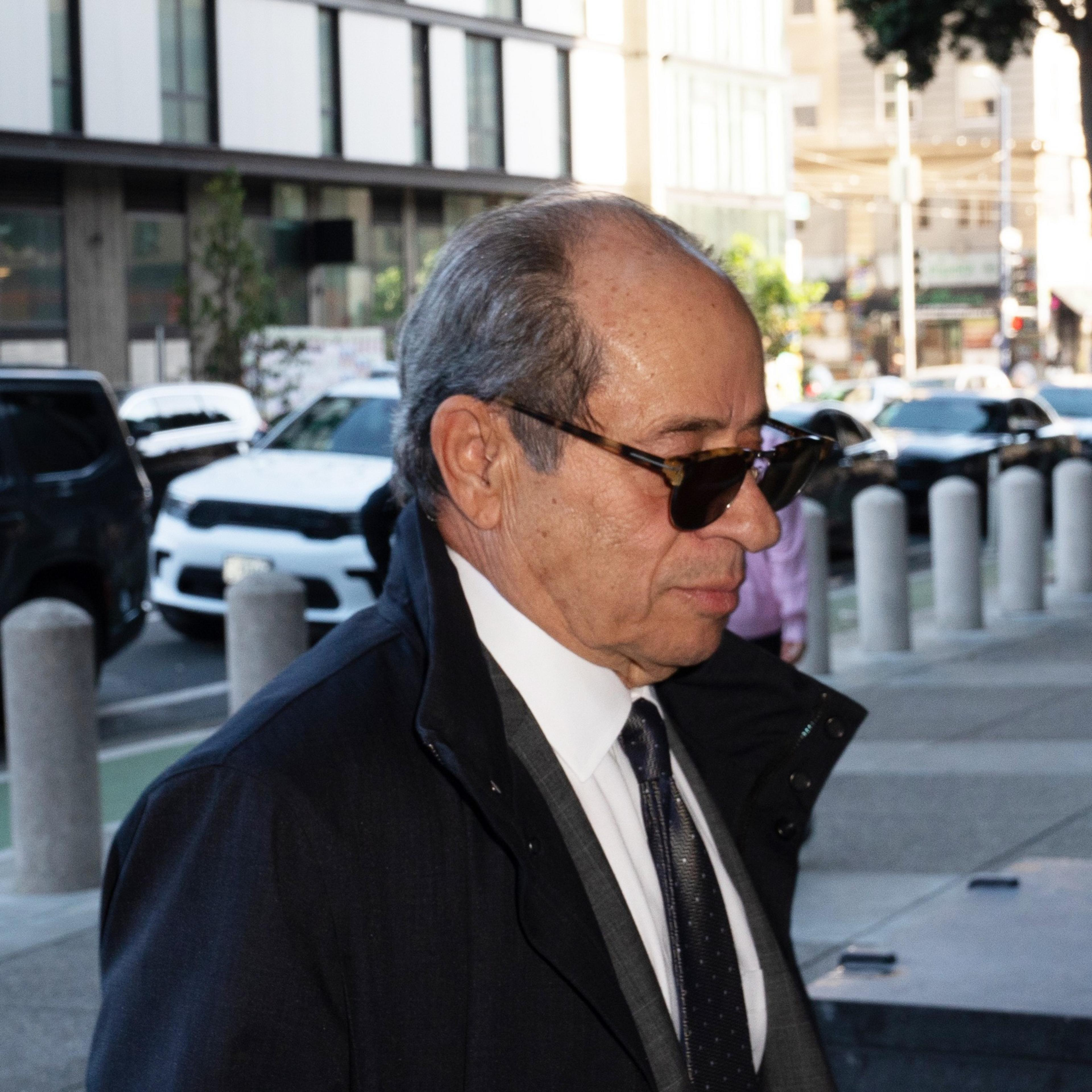 A man in a dark suit and sunglasses walks outside, with cars and buildings in the background.
