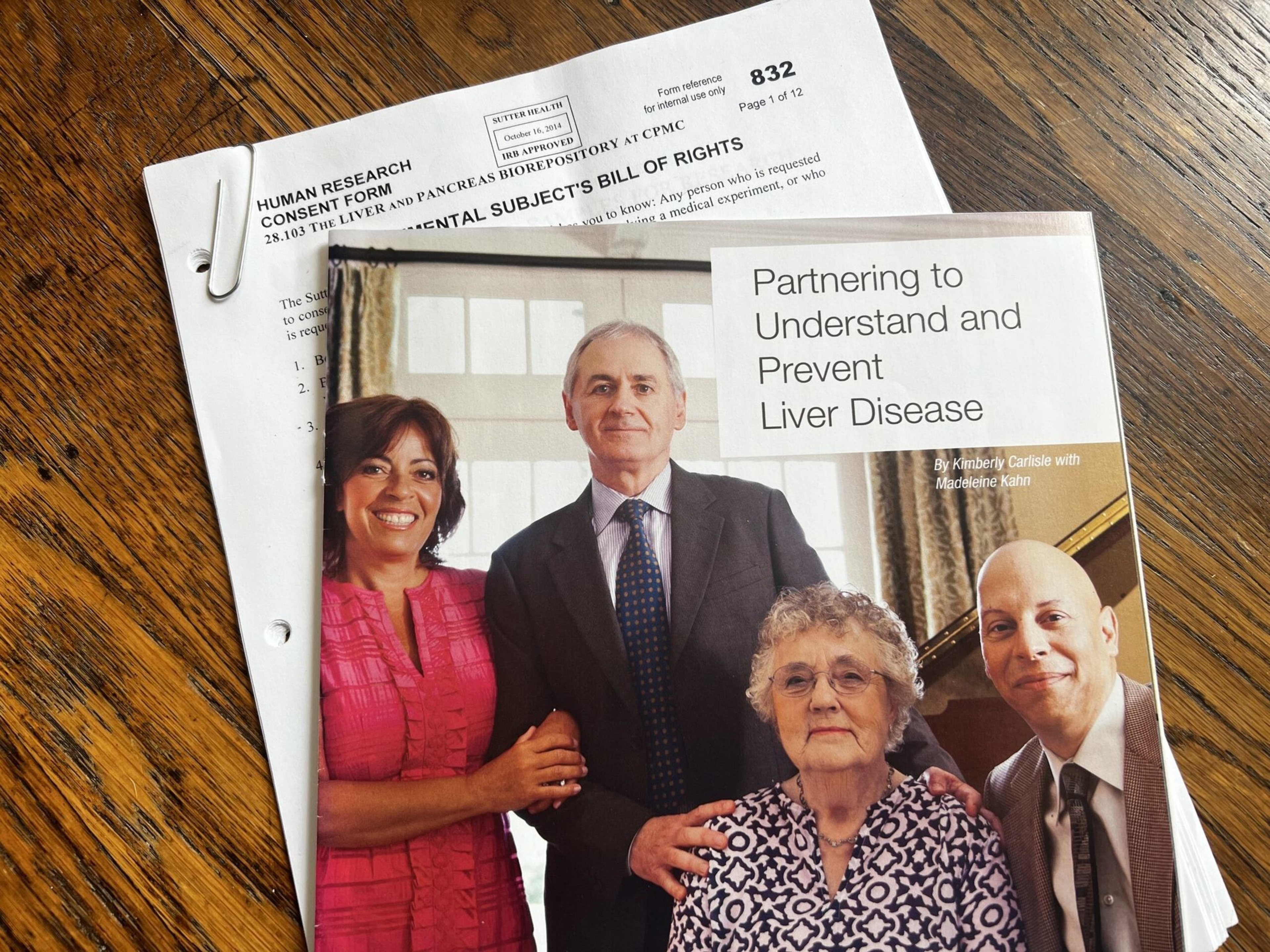 A brochure titled "Partnering to Understand and Prevent Liver Disease" with four people posing together lies on top of a human research consent form on a wooden surface.