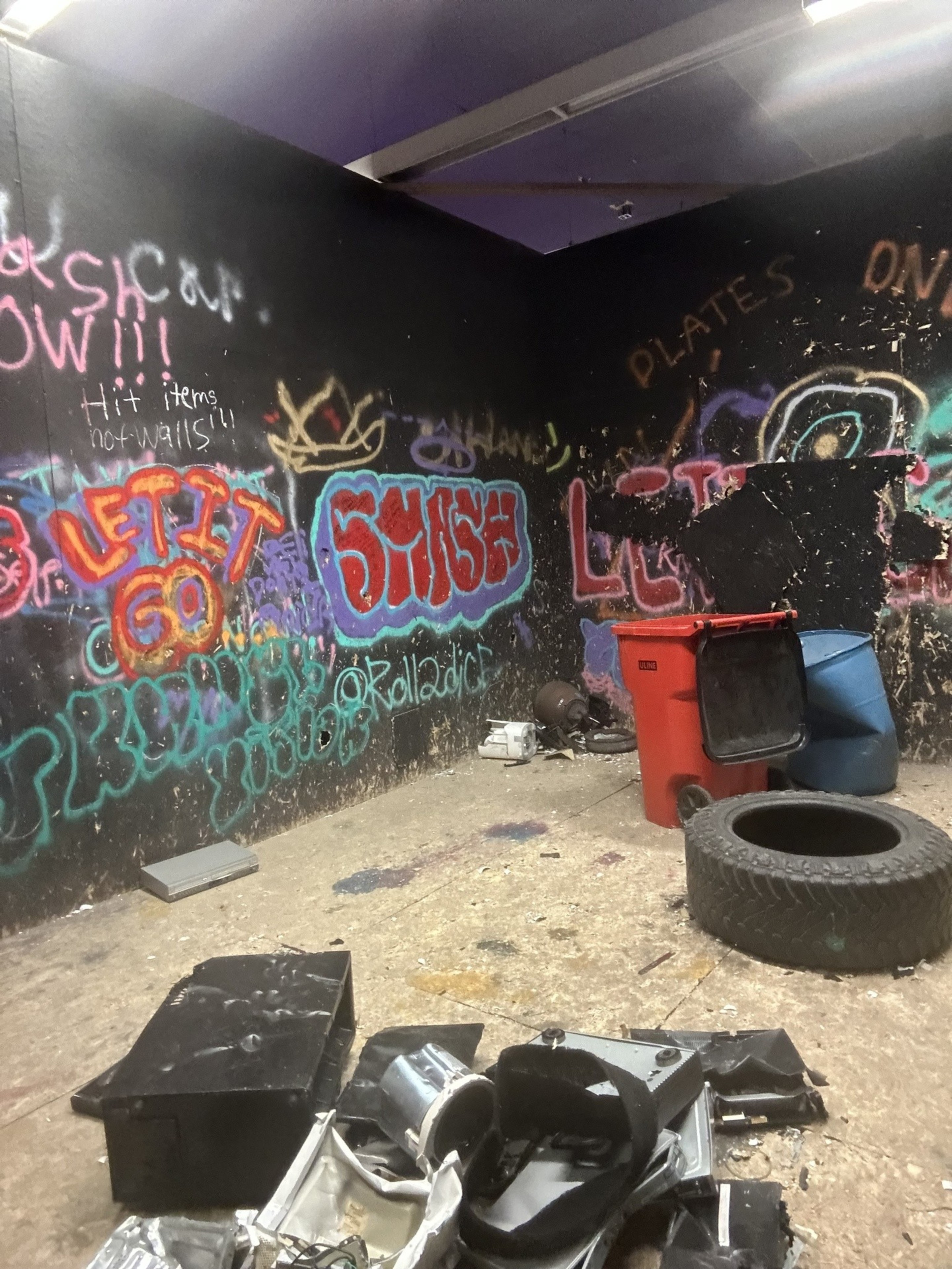 The image shows a dimly lit room with black walls covered in colorful graffiti. The floor is littered with debris, a tire, and broken items. A red bin stands nearby.
