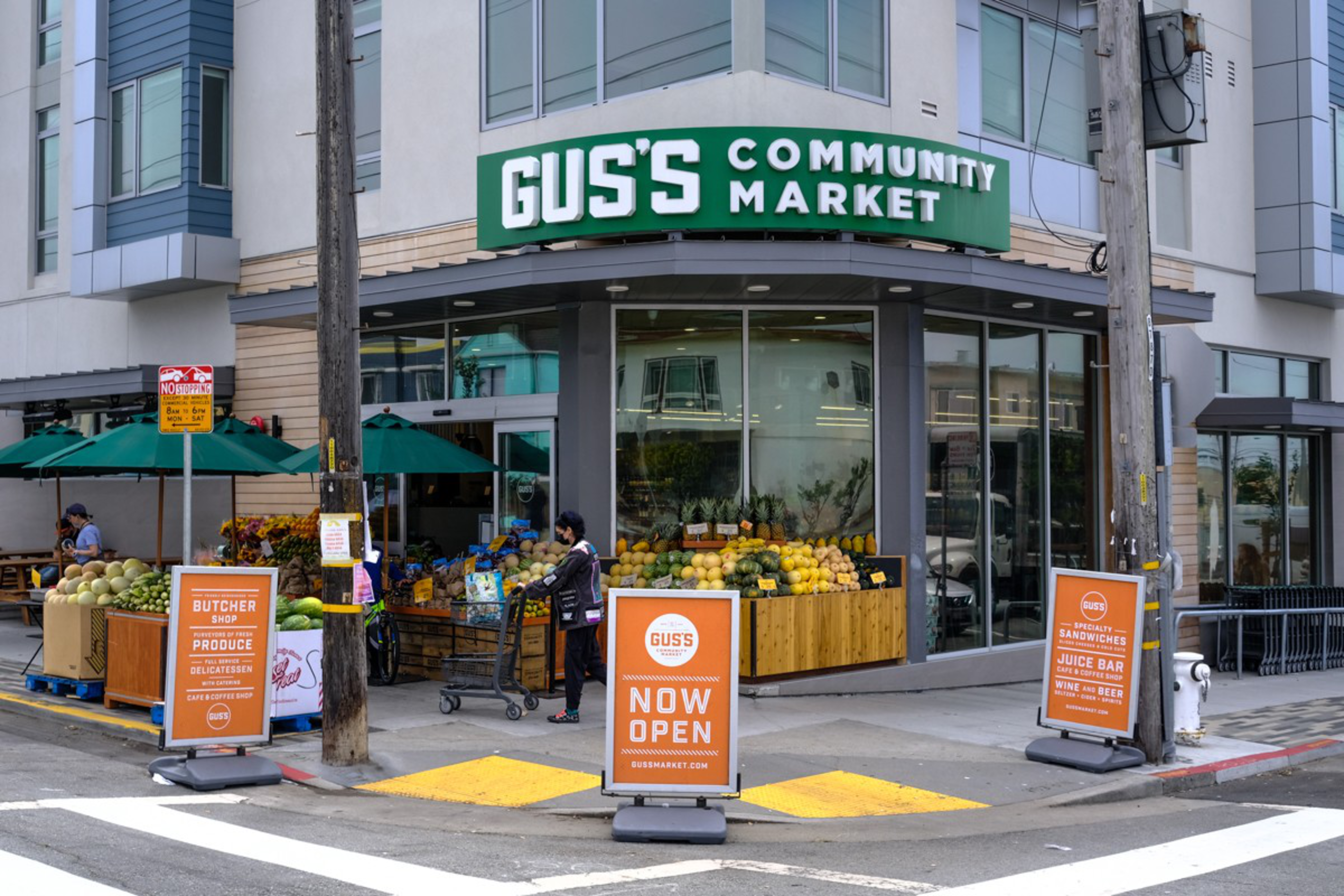 Gus's Community Market storefront displays fresh produce outside with signs announcing it is now open, located at a street corner.