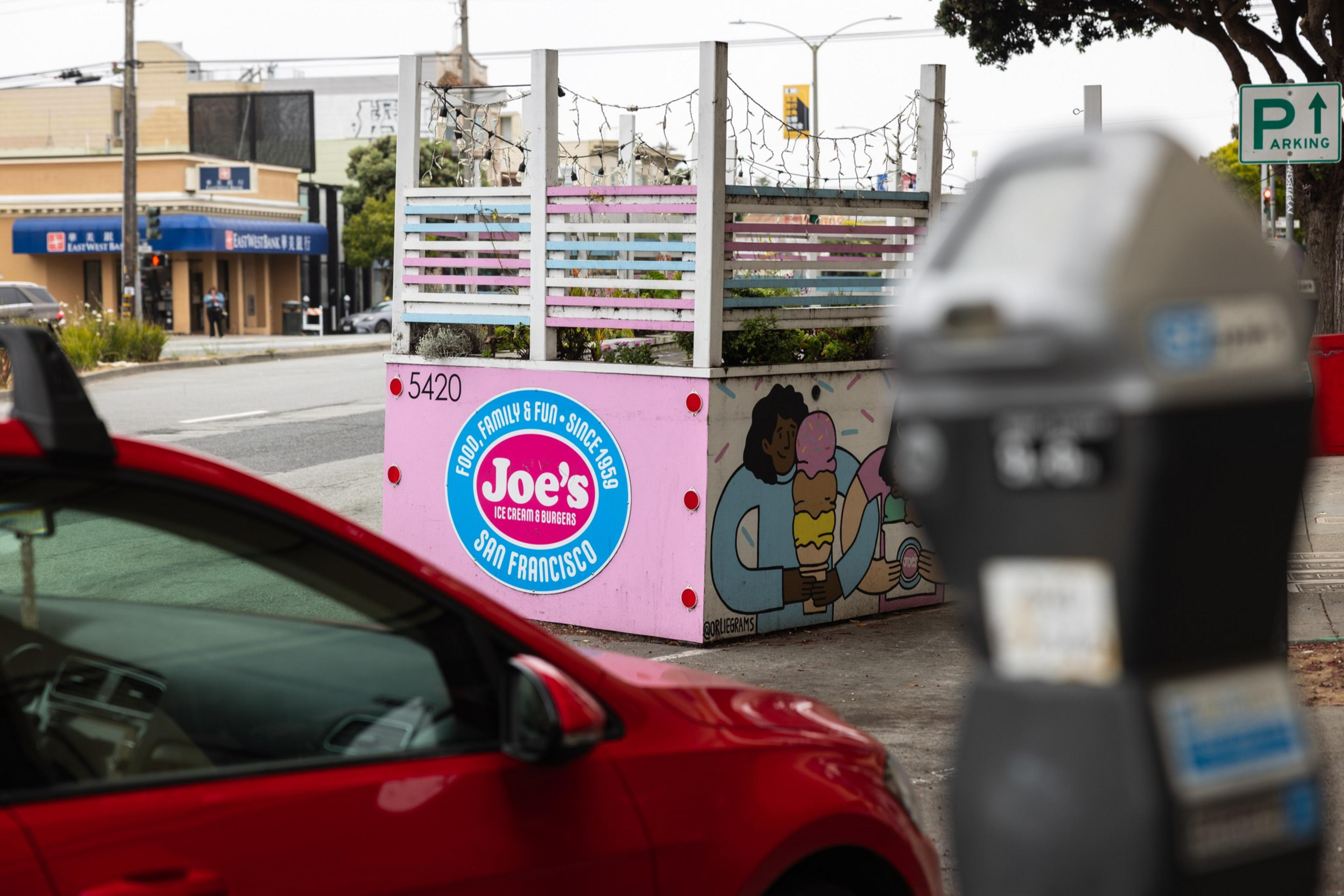 A pink planter box with Joe's Ice Cream & Burgers logo and an illustration of a person holding an ice cream cone sits near a street, with a red car and parking meter visible.