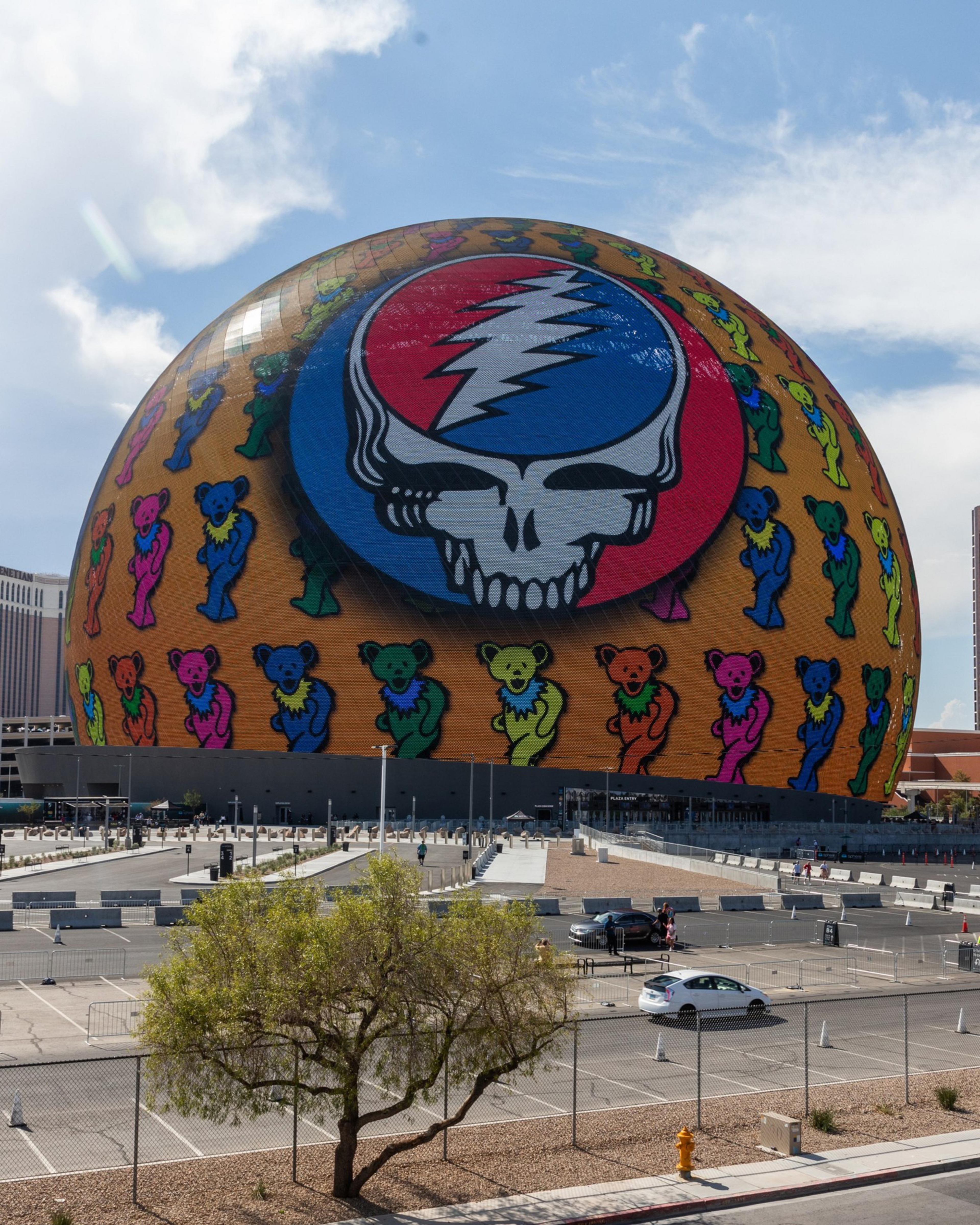 A large, round building displays a colorful graphic featuring a skull with a lightning bolt and dancing bears. The foreground includes a parking lot and a tree.