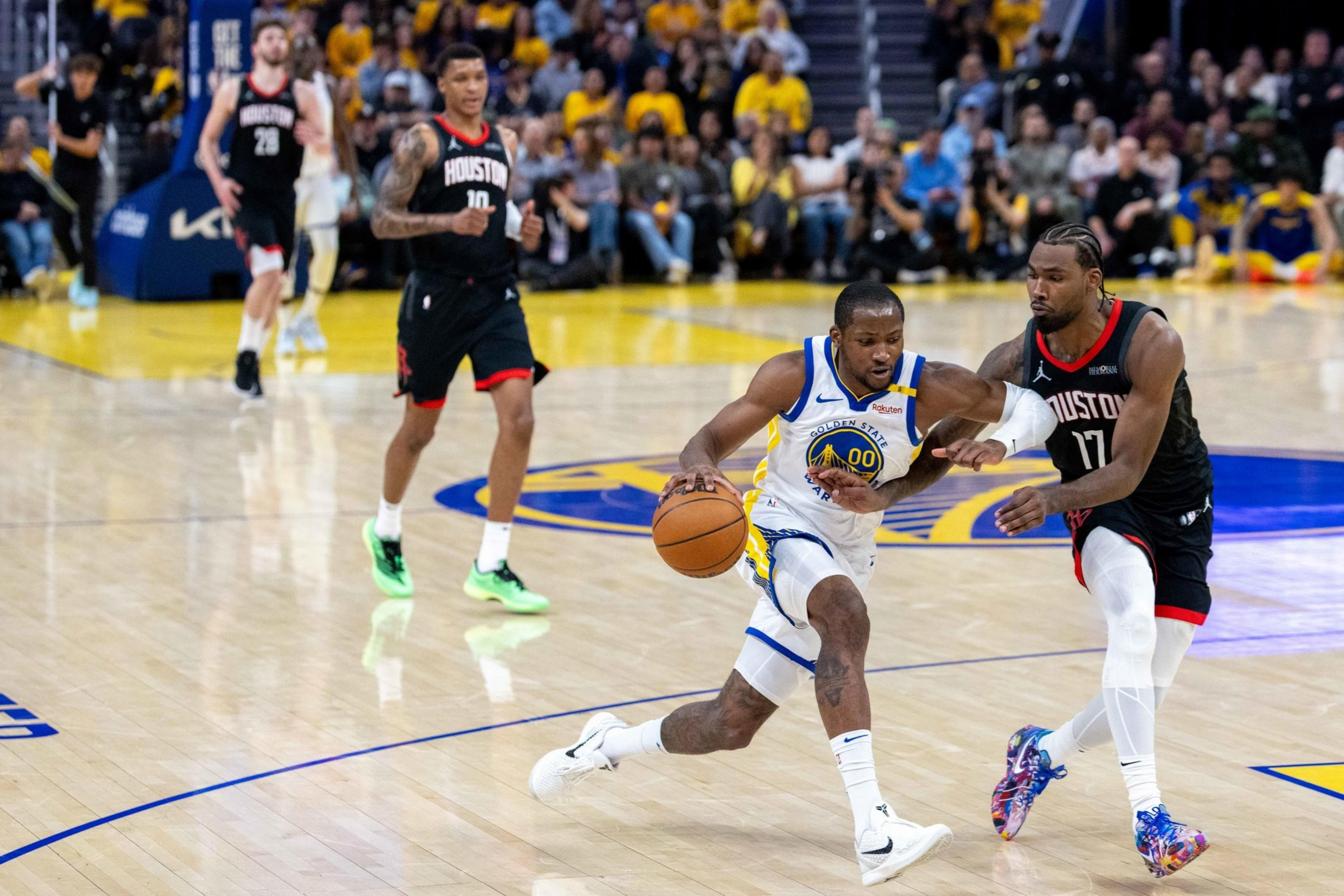 A basketball player in a white uniform drives past a defender in a black uniform on a court with a crowd in the background.
