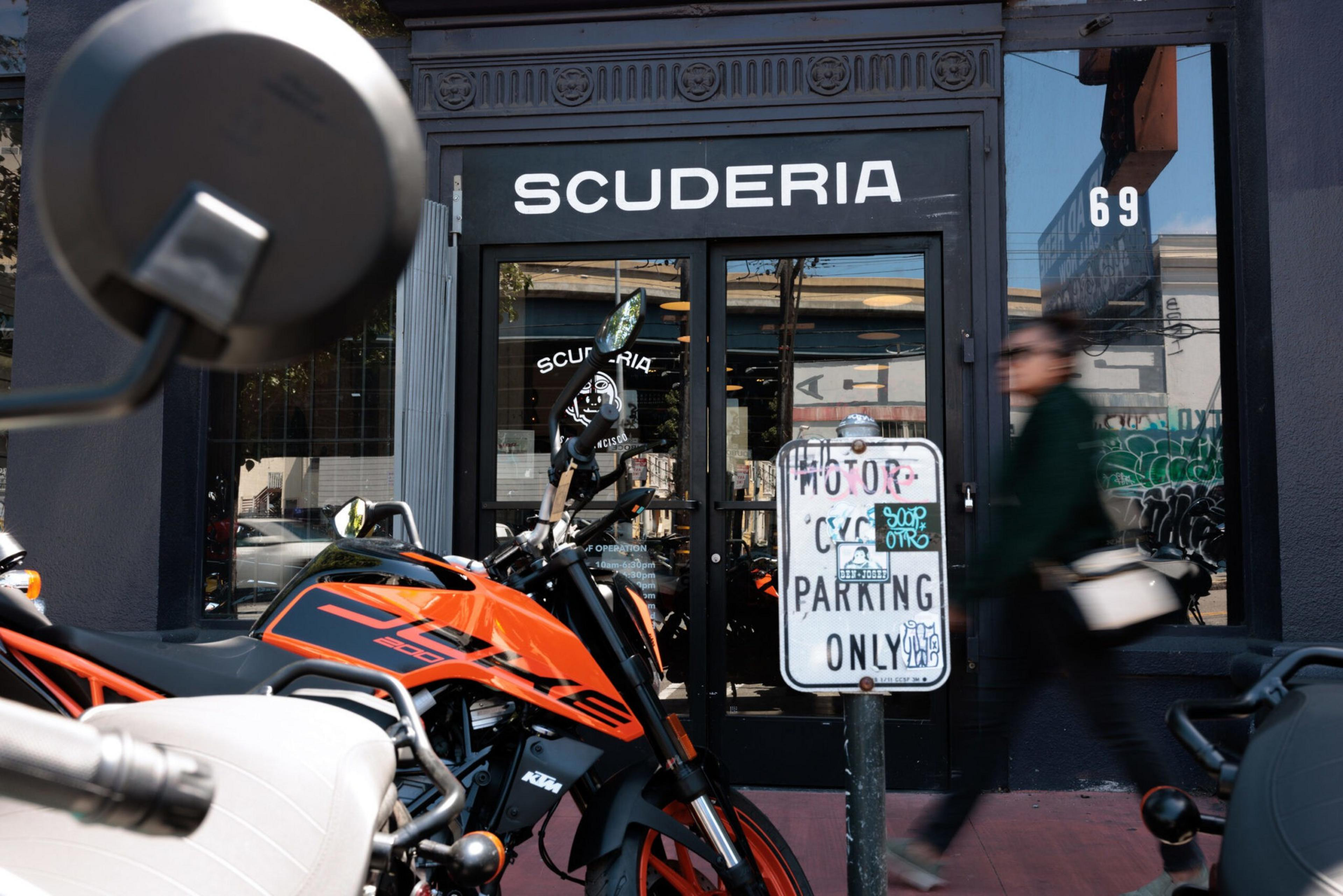 A black and orange KTM motorcycle is parked outside a storefront labeled "SCUDERIA" with a sign that reads "MOTORCYCLE PARKING ONLY," while a person walks by.
