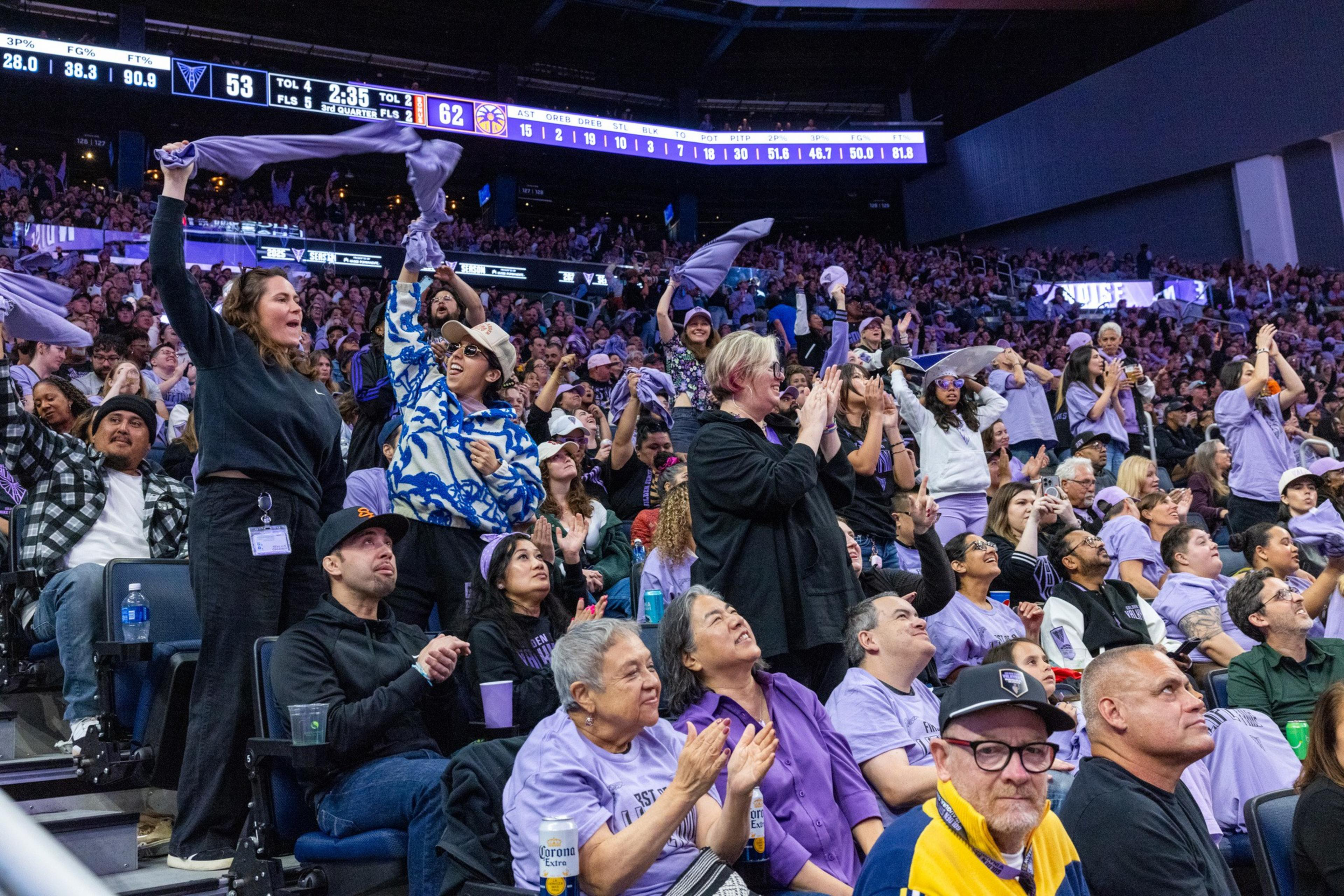 The image shows a lively crowd at a sports game, with fans cheering and waving towels. People are dressed in purple and some are clapping enthusiastically.