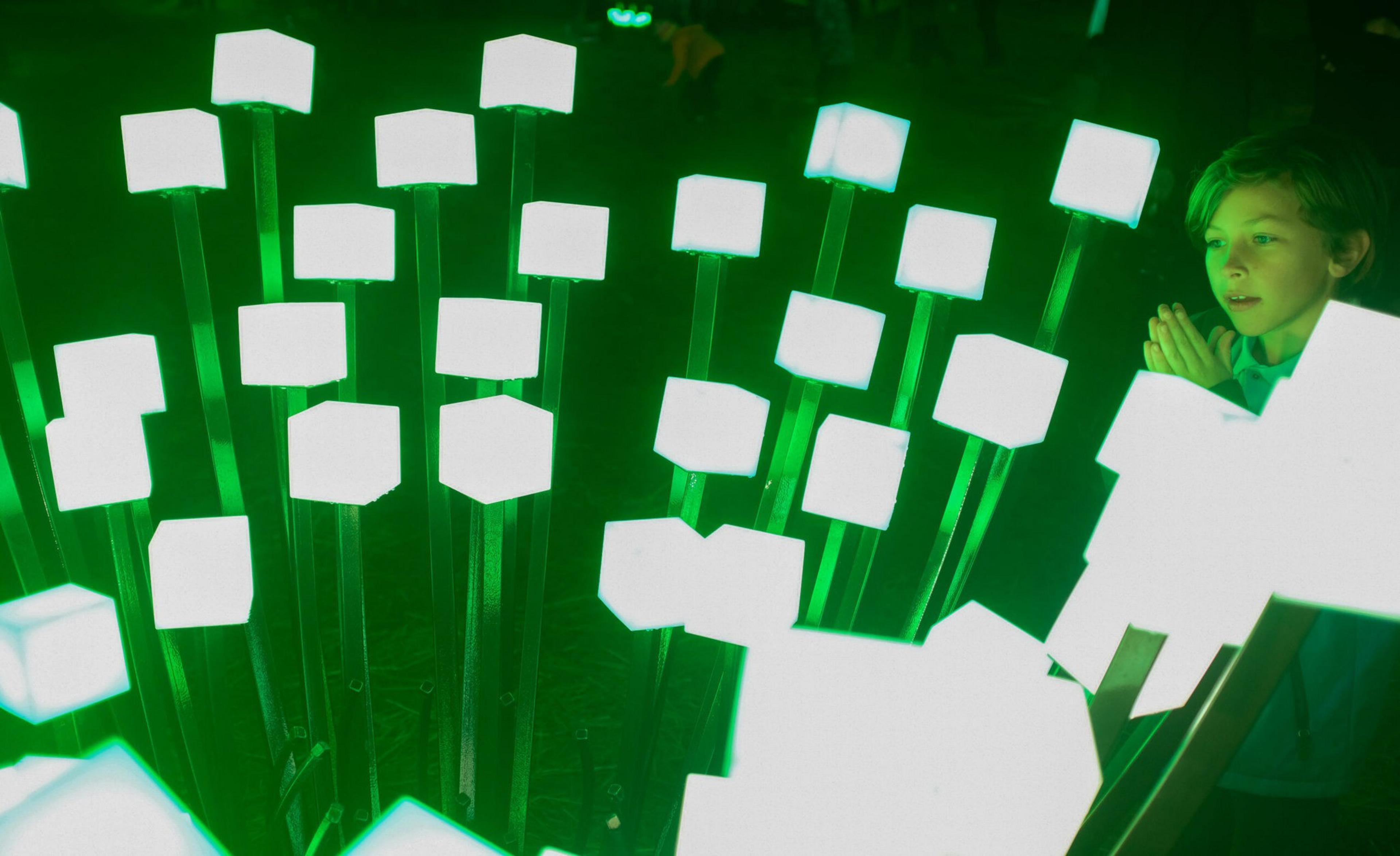 A child watches glowing white cubes mounted on green-lit vertical poles in a dark environment, appearing curious and focused.