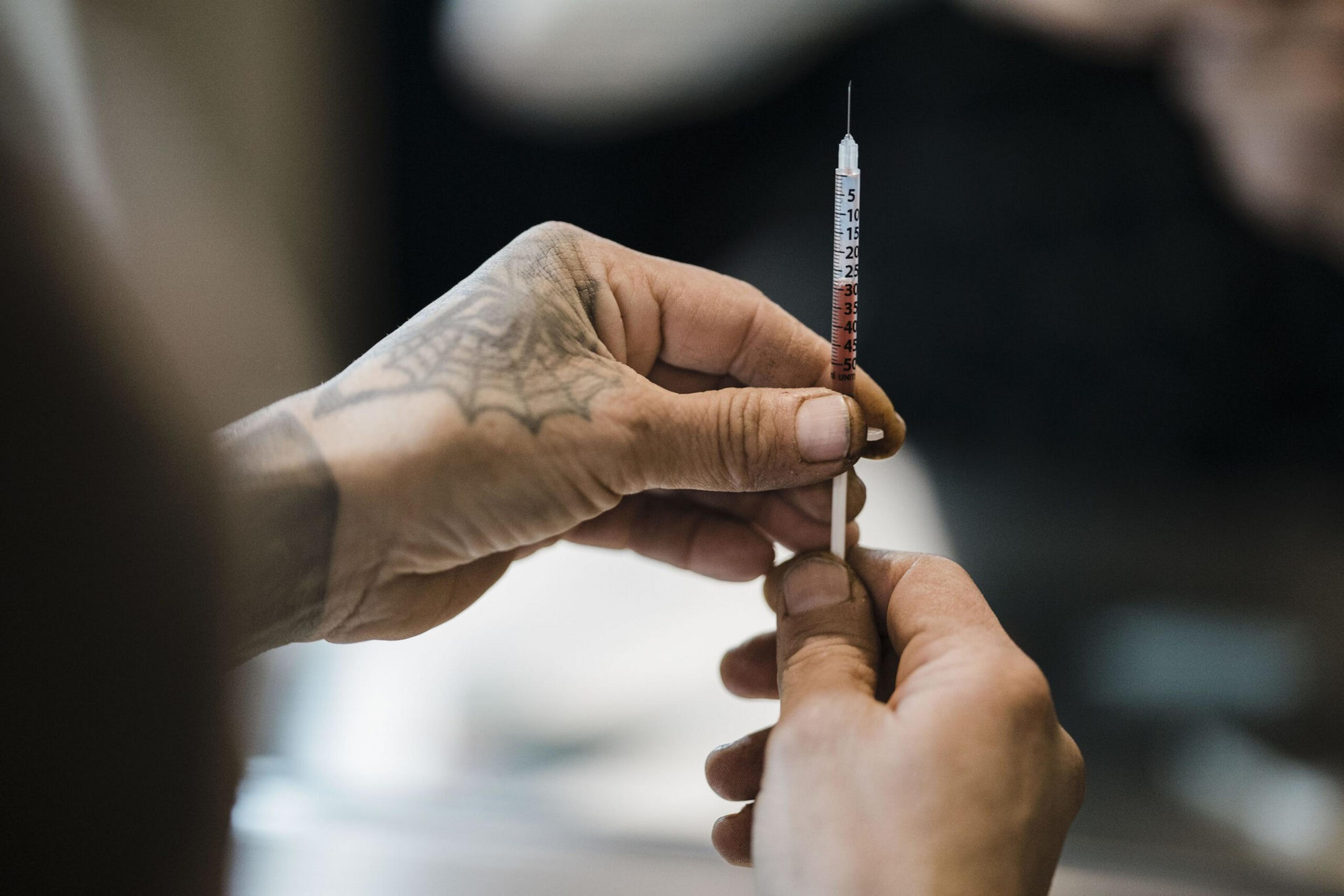 A tattooed hand holds a syringe filled with red liquid, with another hand steadying the syringe below the needle in a close-up shot.