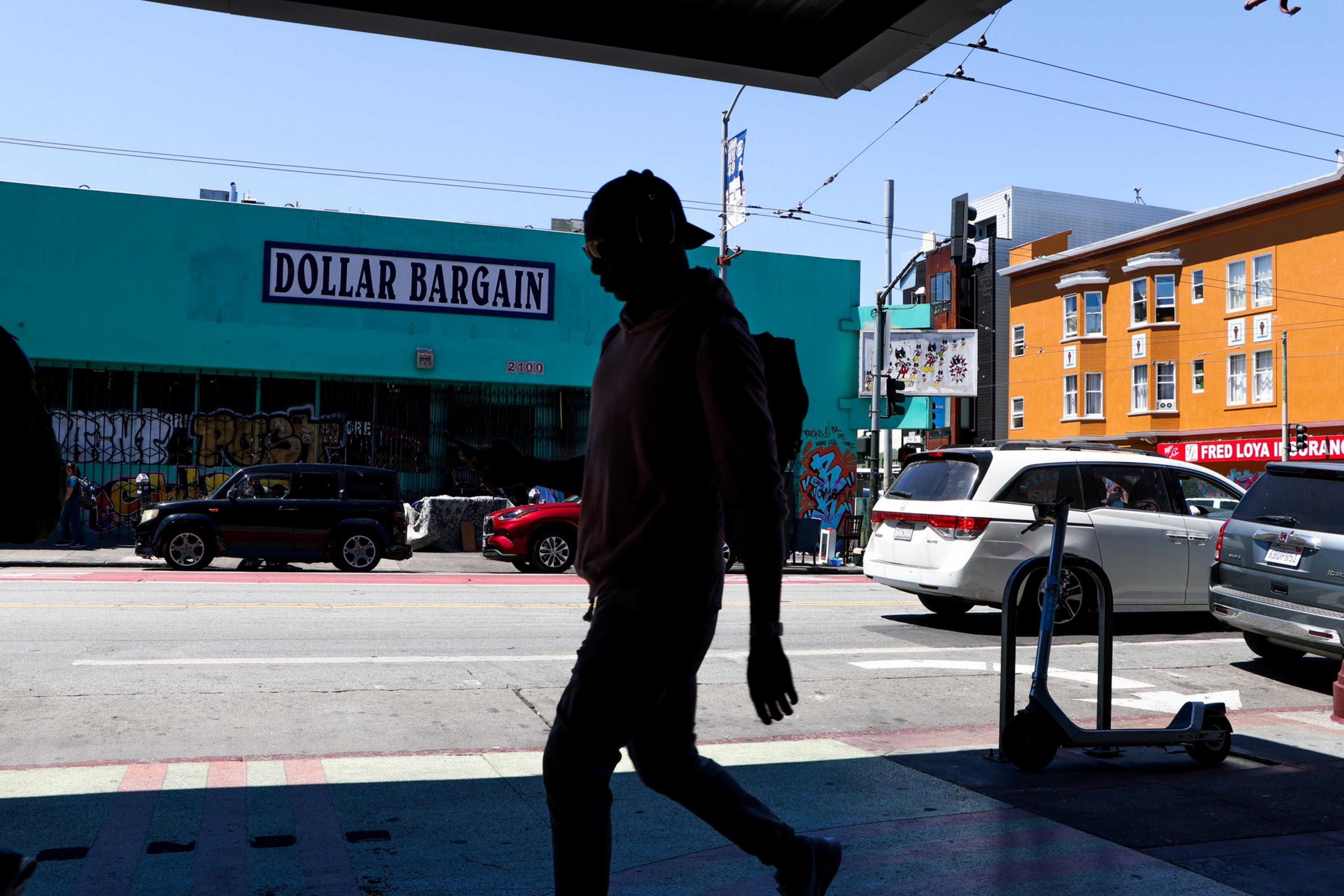 A silhouette of a person walking past a street with parked cars and a store named "Dollar Bargain" on the opposite side under a clear blue sky.