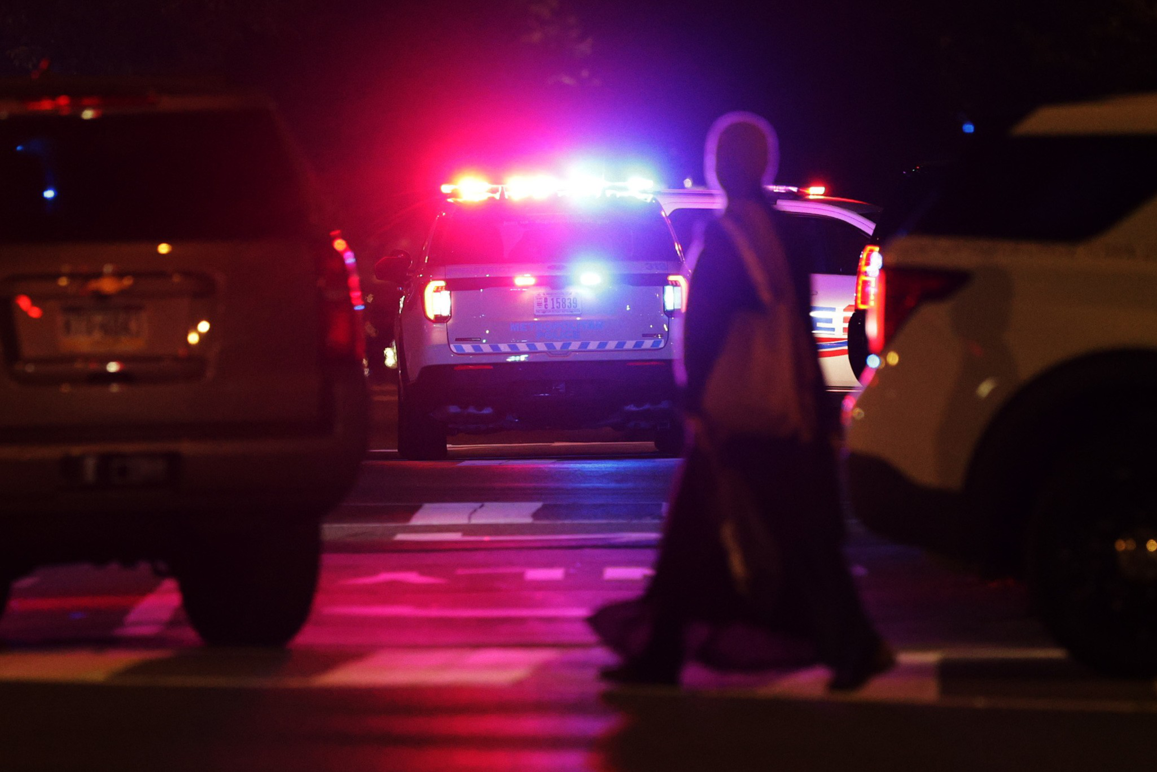 A person walks across a street at night near parked cars, while a police vehicle with bright red and blue flashing lights is stopped ahead.