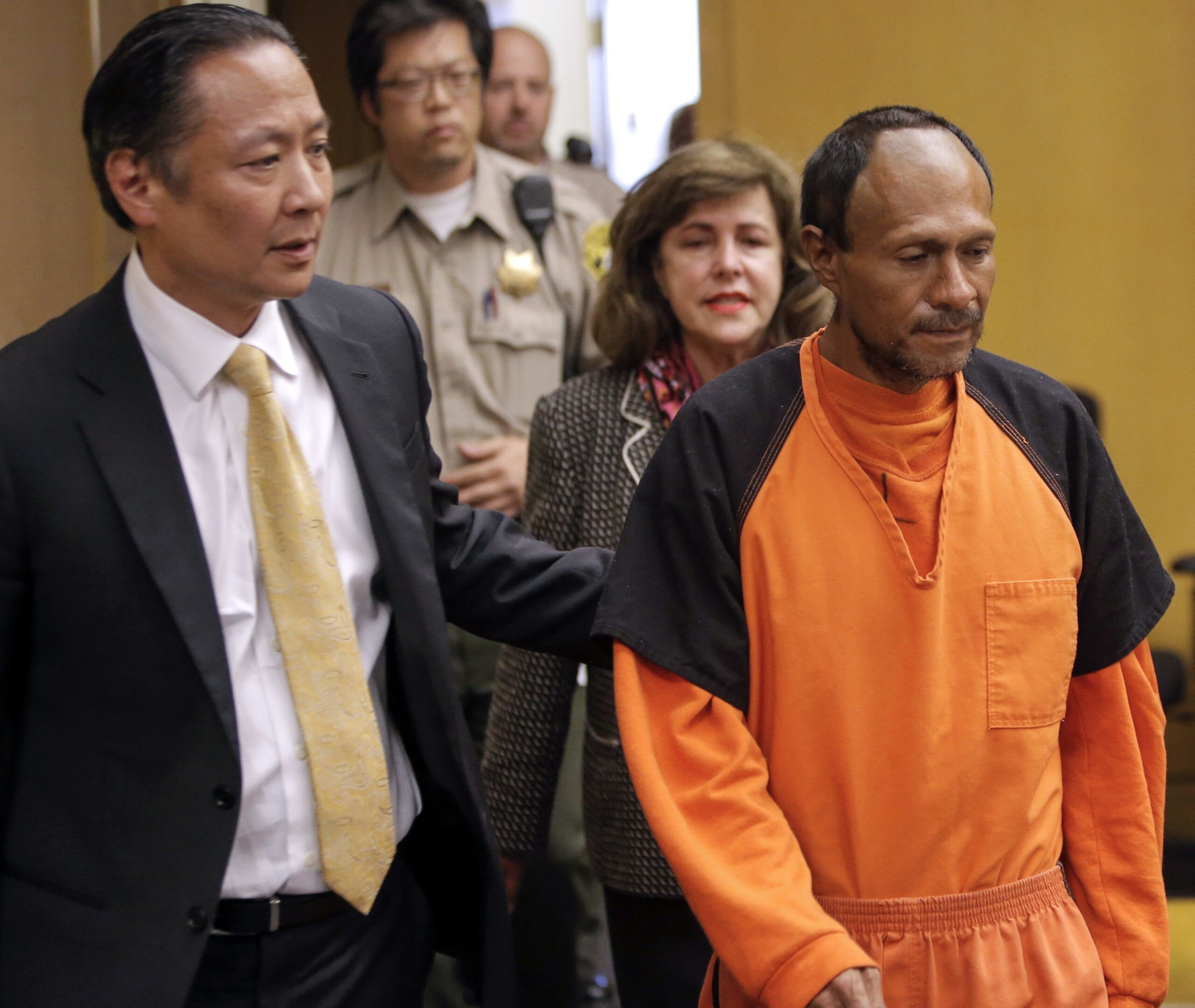 A man in an orange prison jumpsuit is escorted by a suited man, with two other officials, including a uniformed officer, behind them in a hallway.