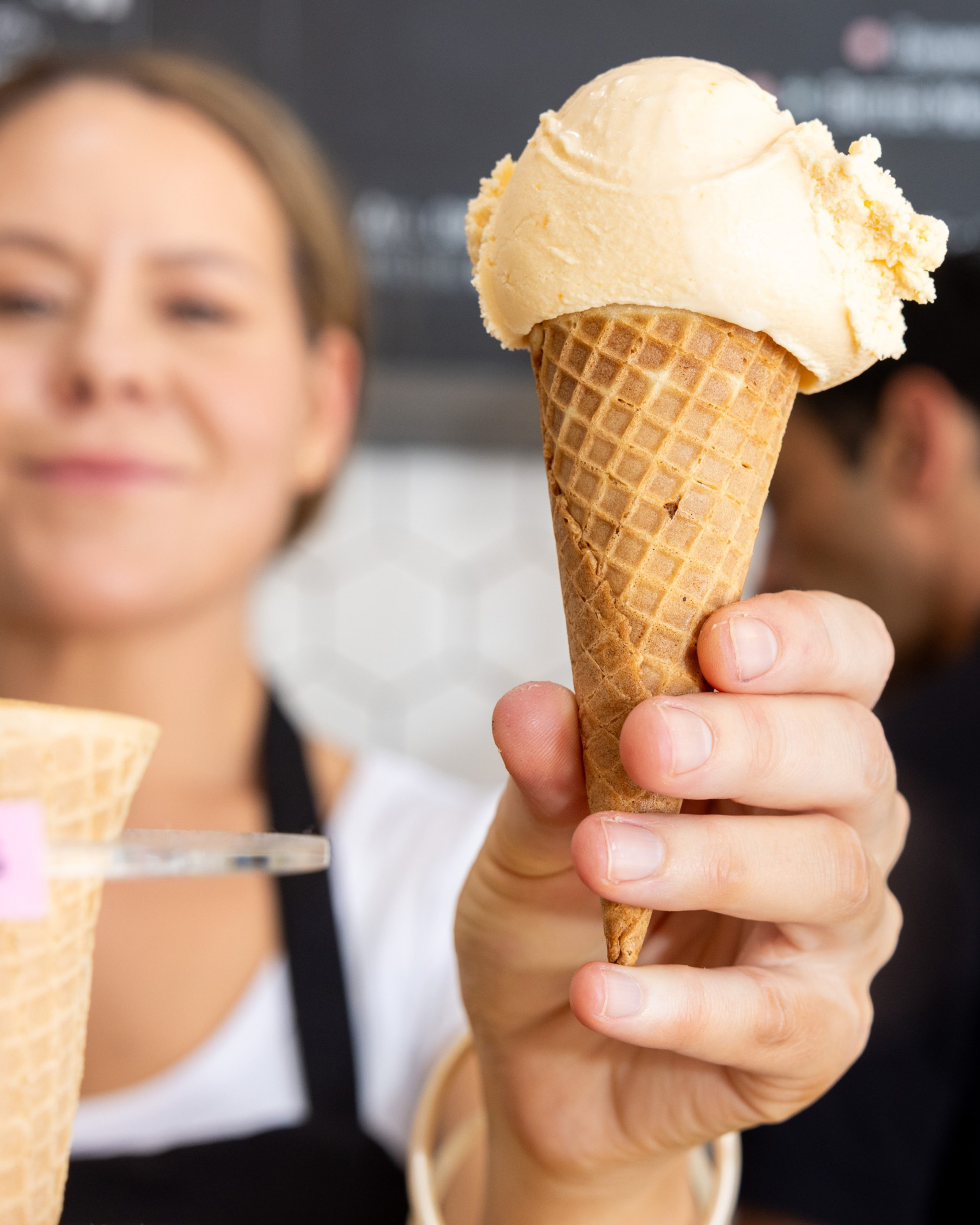 A person wearing a black apron holds a waffle cone with vanilla ice cream in focus, with their face out of focus in the background.