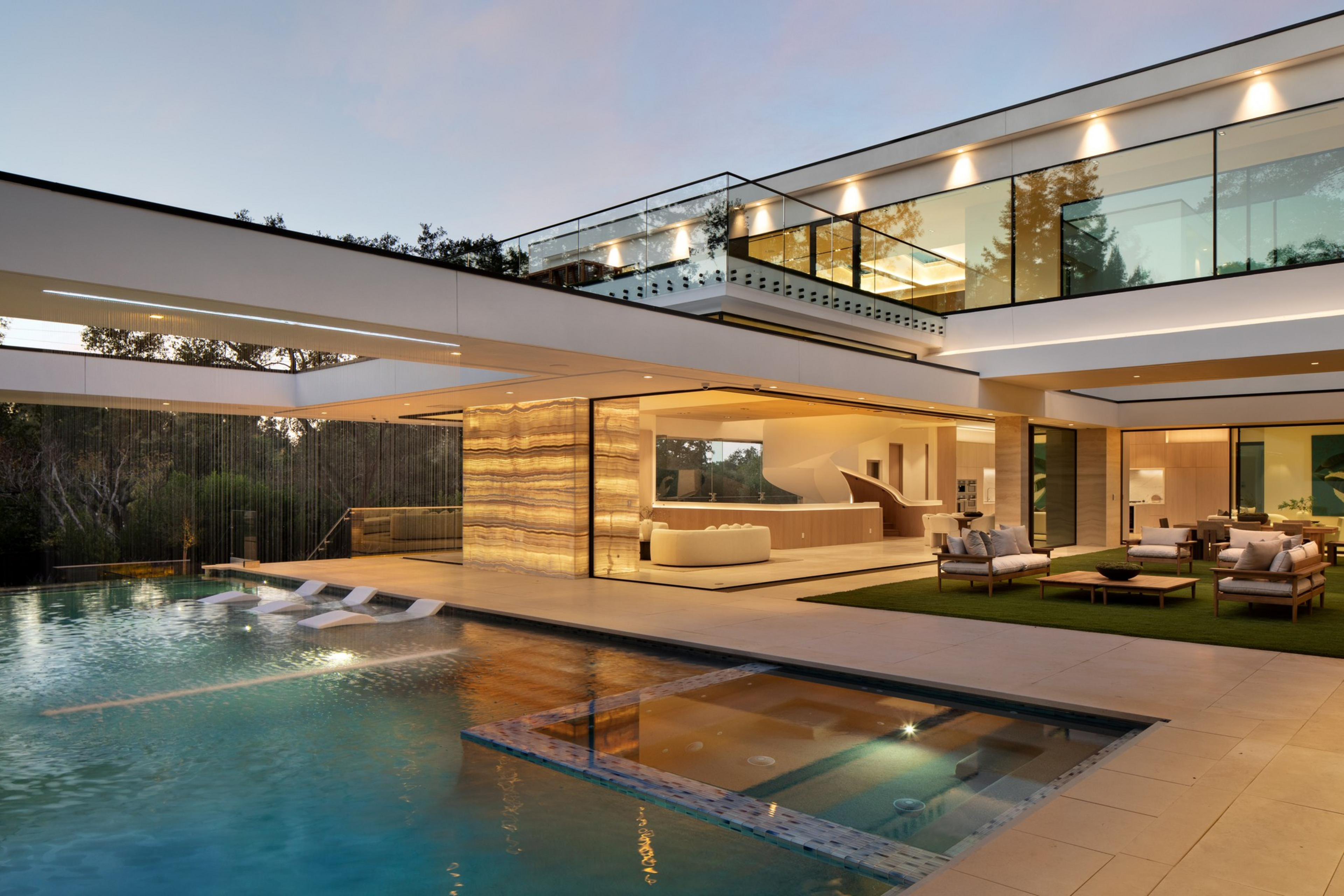 A modern luxury house features an open living area, glass railing on the upper floor, and a spacious pool with built-in loungers and a hot tub.