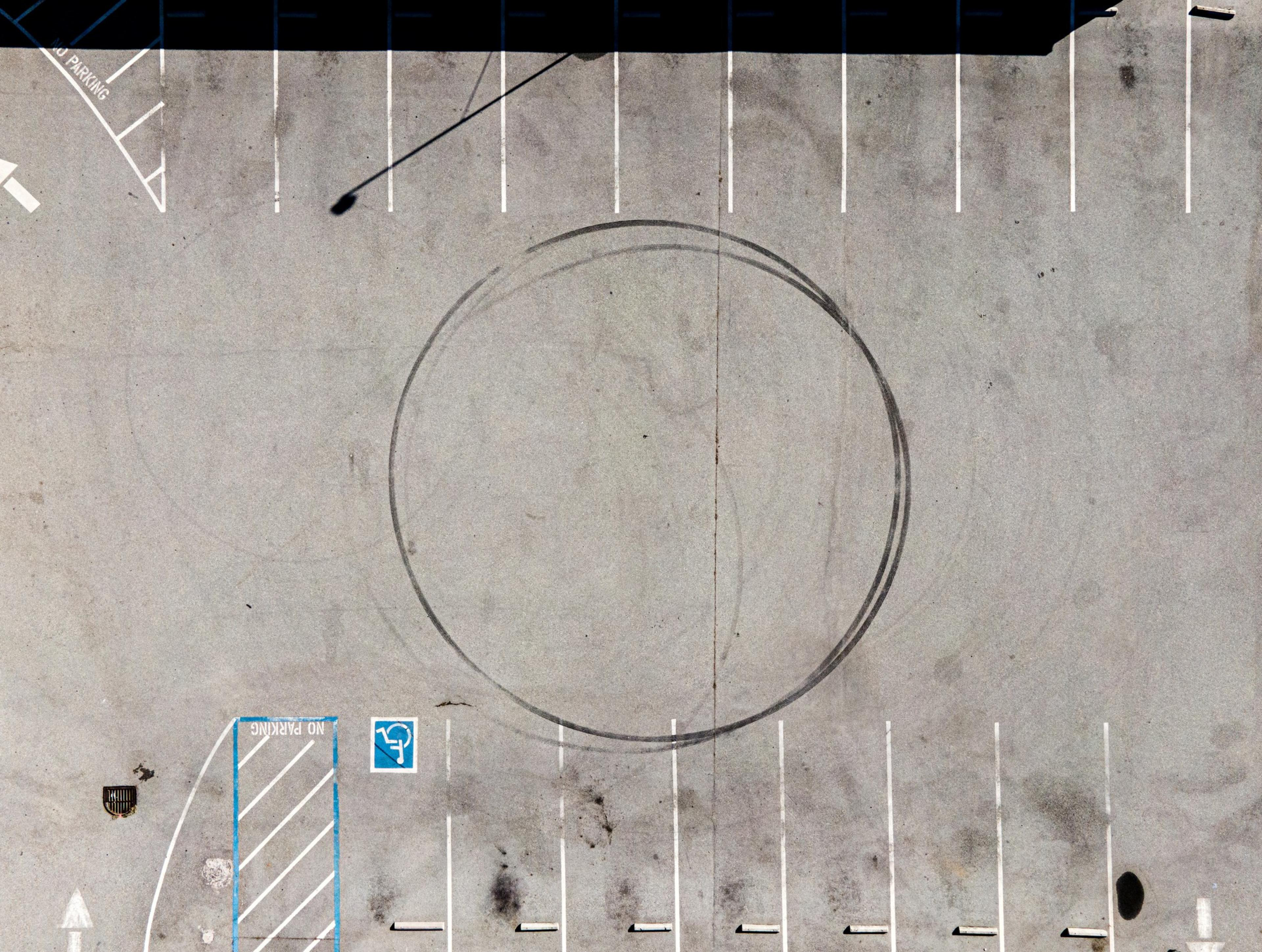 Aerial view of an empty parking lot with a large circular tire skid mark in the middle, numerous white parking lines, and a marked blue handicap spot.