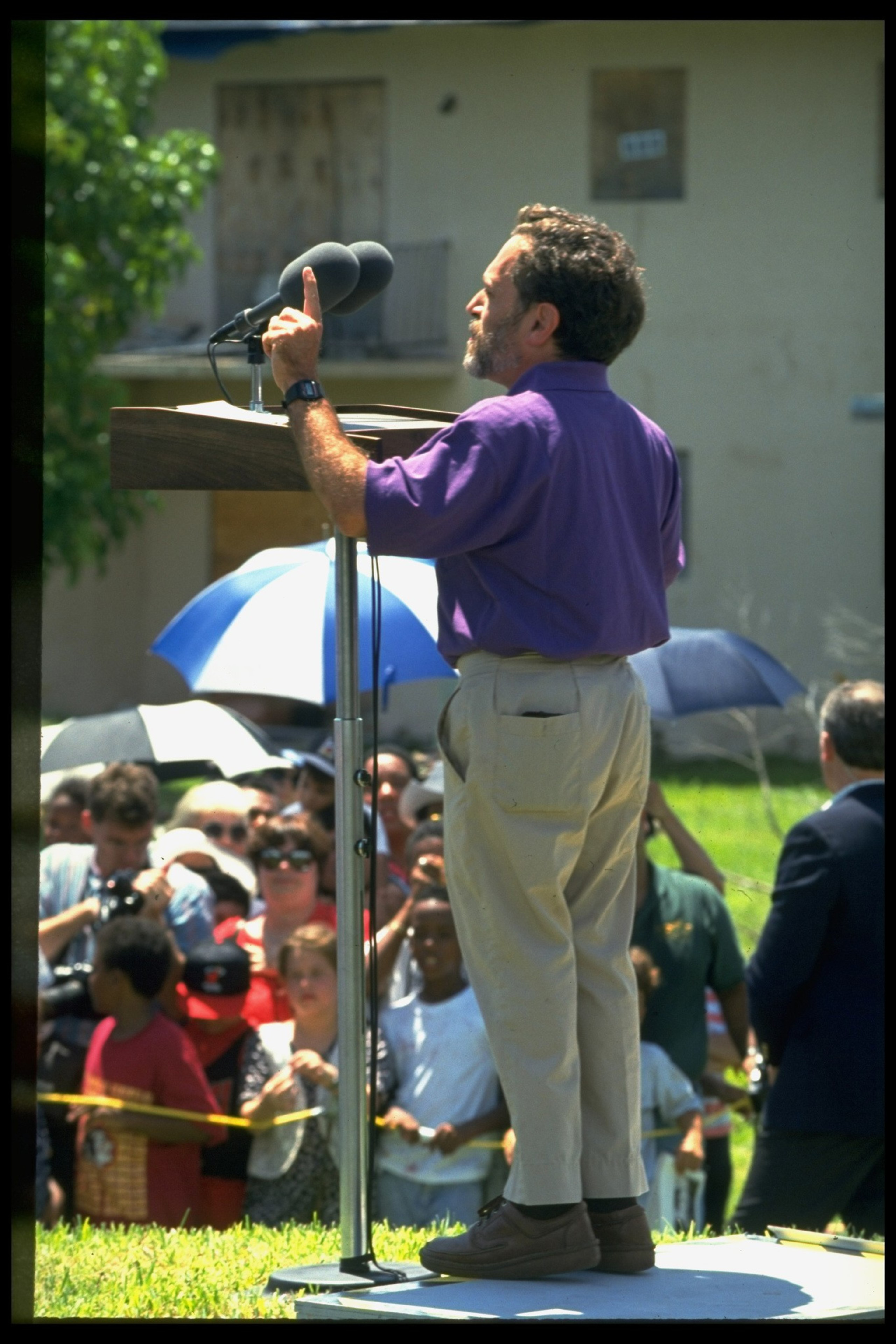 A man in a purple shirt and beige pants speaks passionately at a podium to a crowd, some holding umbrellas for shade.