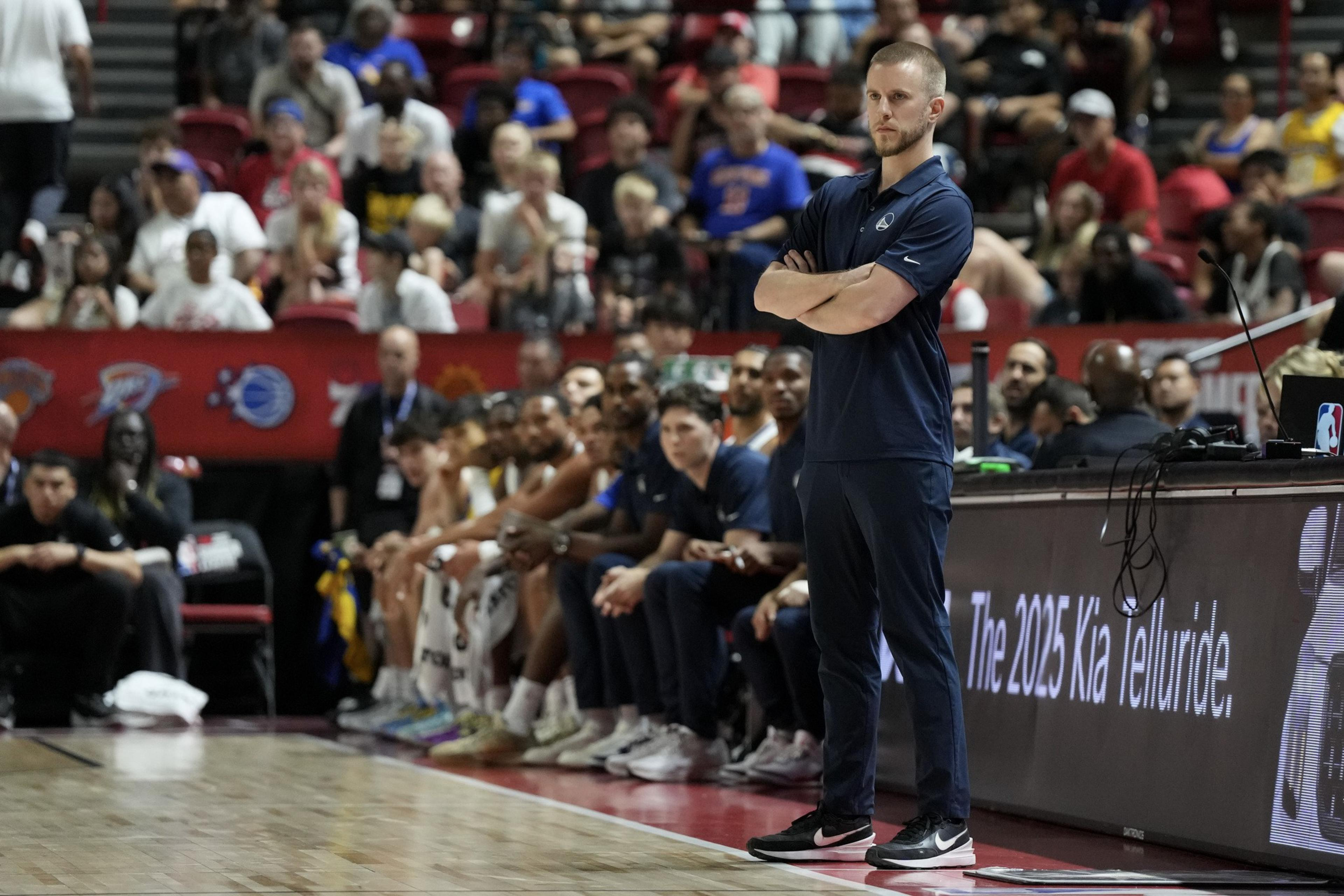 A coach stands with arms crossed on a basketball court's sideline, while players sit on the bench and an audience watches intently.