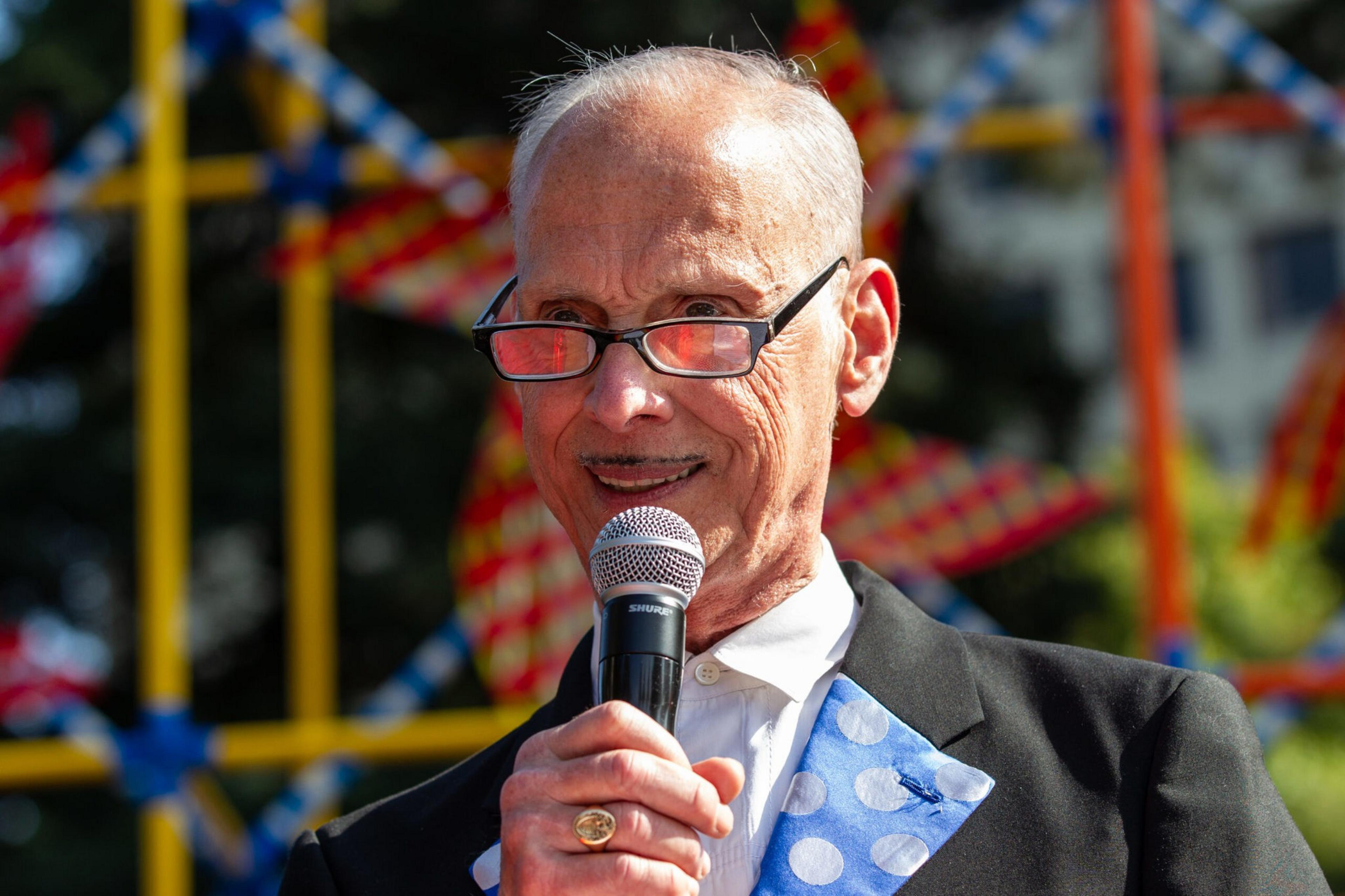 An elderly man with glasses holds a microphone and smiles, wearing a suit with a polka-dot blue vest and tie behind a colorful background.