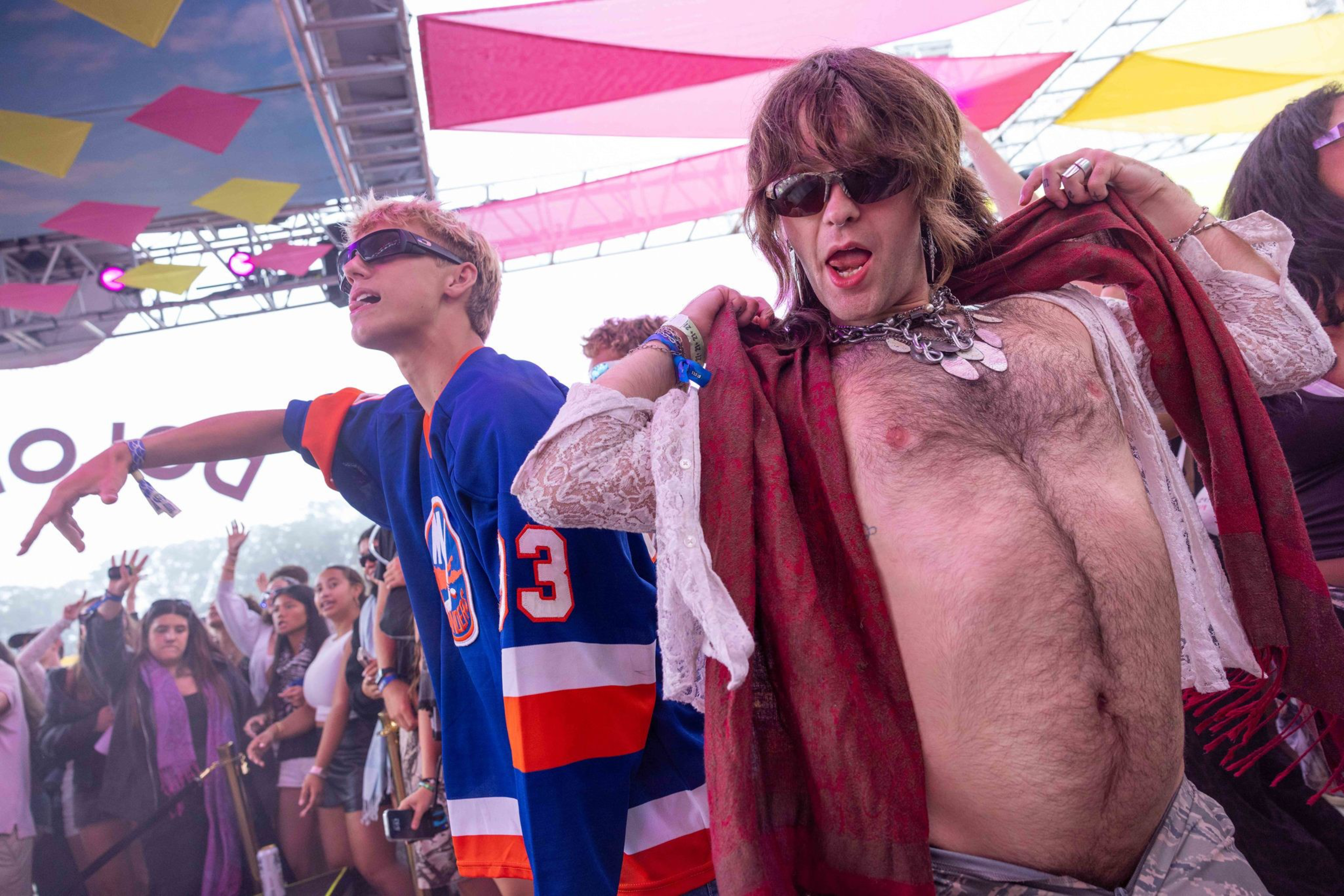 A man with a hairy chest poses confidently wearing sunglasses, a lace shirt, and a red scarf, while a young man in a blue sports jersey dances nearby at a lively outdoor event.