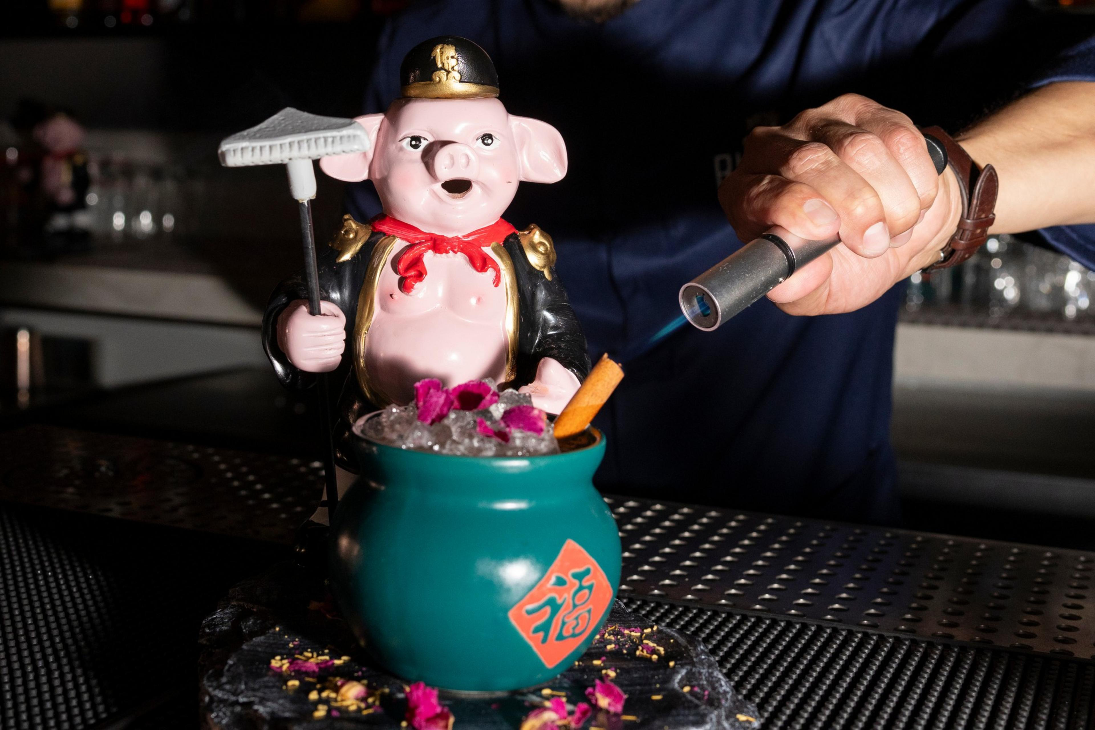 A bartender is torching a drink in a quirky container shaped like a pig dressed as a soldier, holding ice and garnished with flower petals and a cinnamon stick.