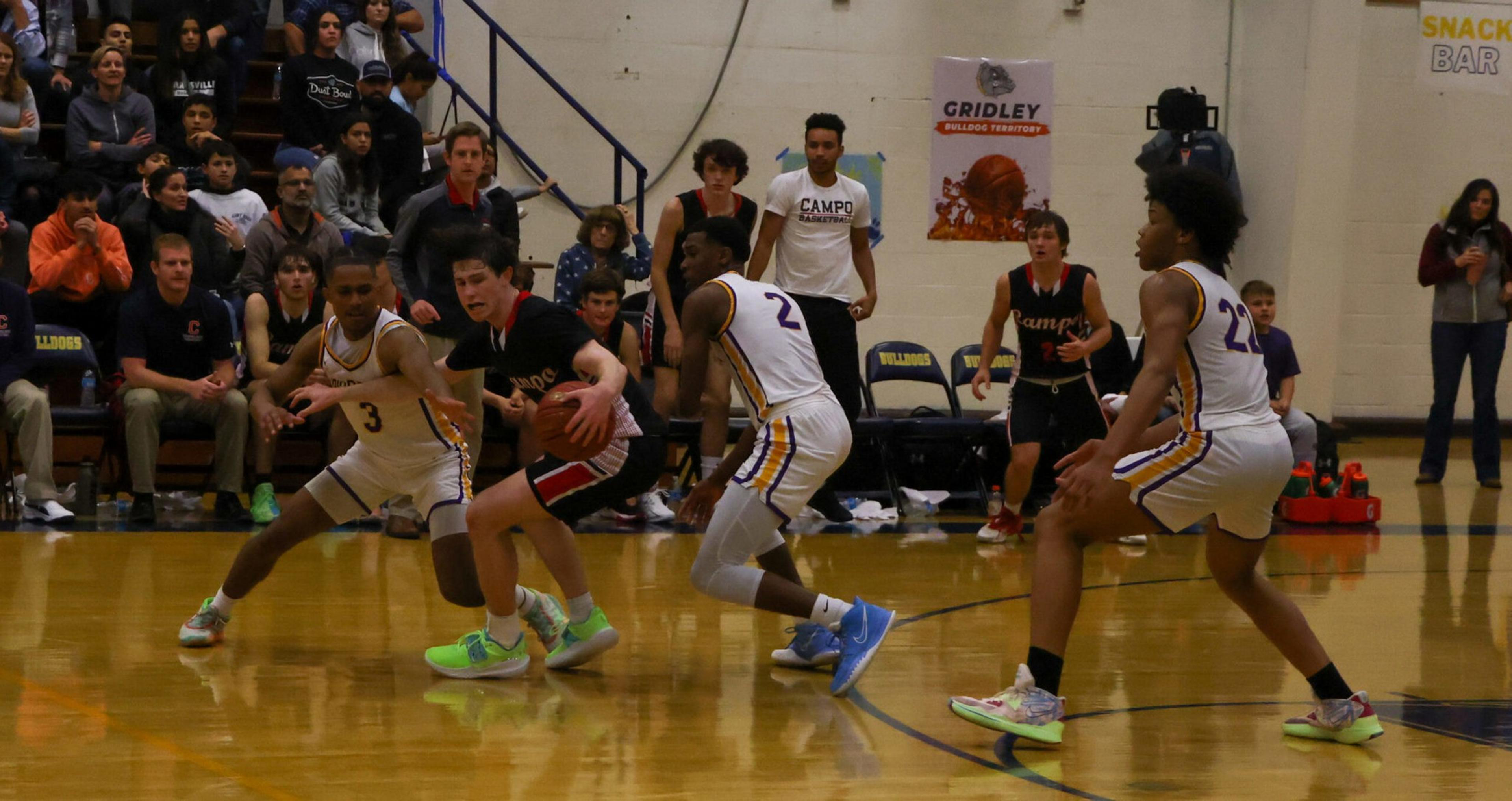A basketball player in a black uniform dribbles while three defenders in white uniforms closely guard him on a gym court.