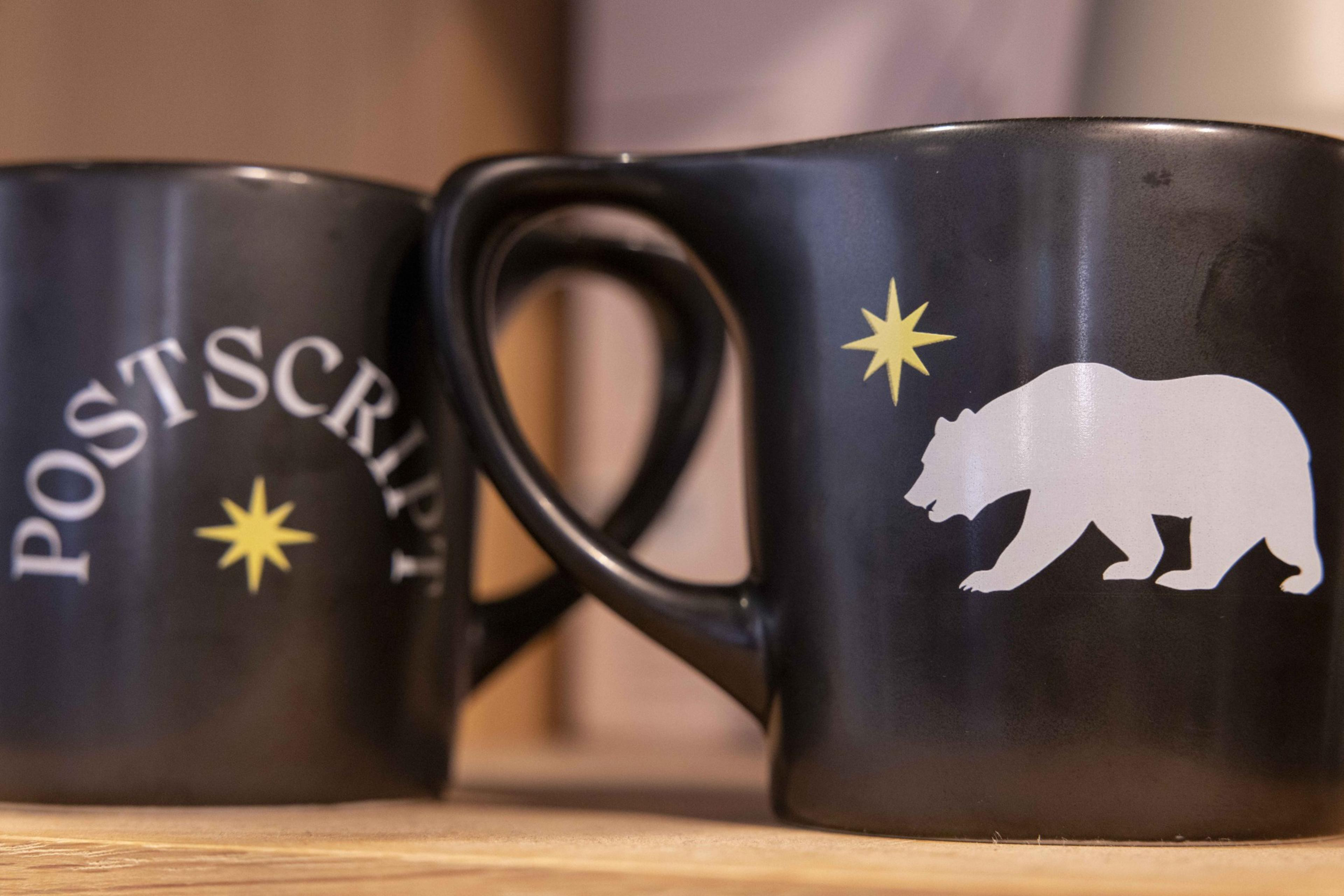 Two black mugs are close together. One displays the word &quot;POSTSCRIPT&quot; and a yellow star, while the other has a white bear and a similar yellow star.