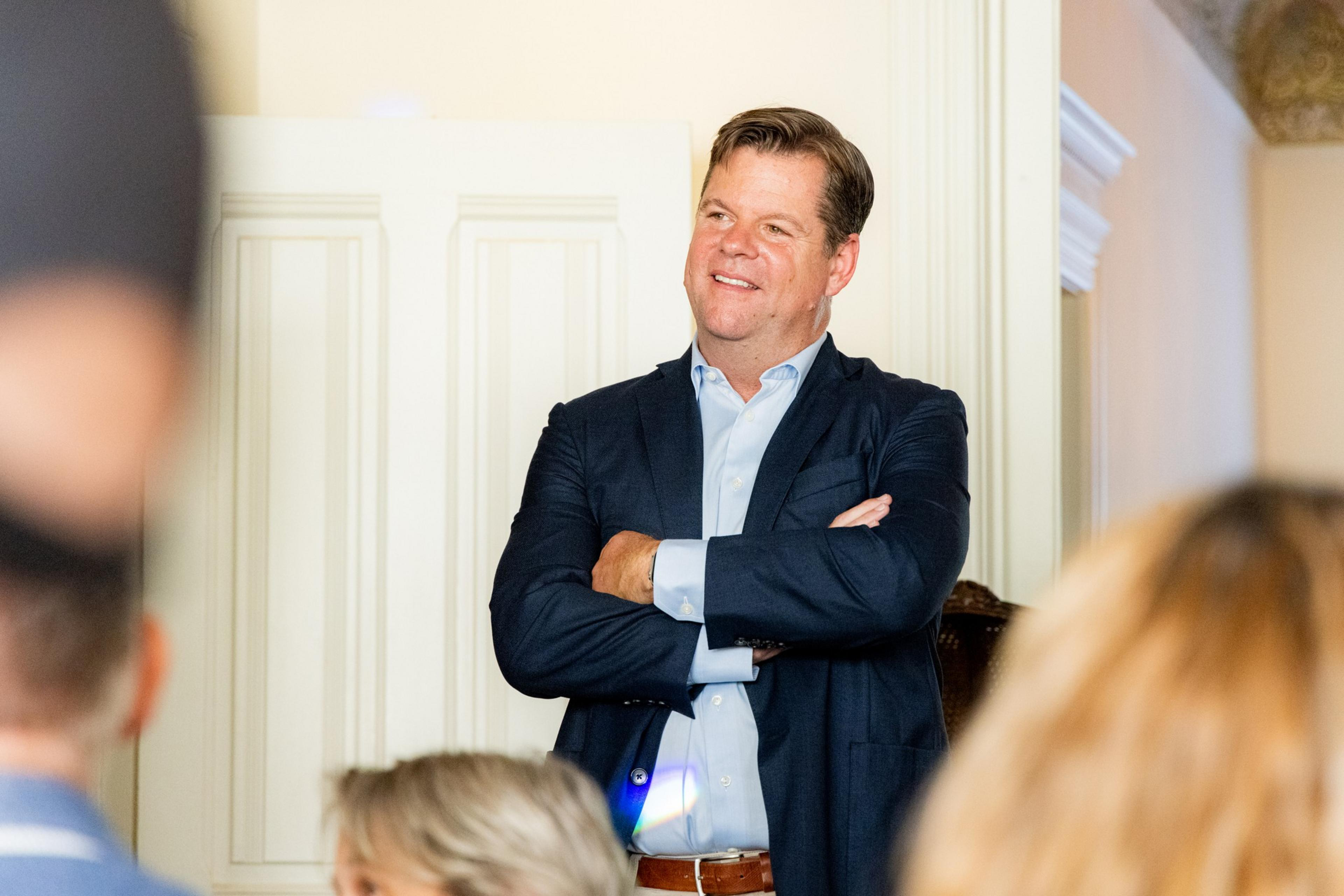 A man in a suit stands smiling with arms crossed. He's surrounded by people, facing them, and appears to be in a formal setting with paneled walls.
