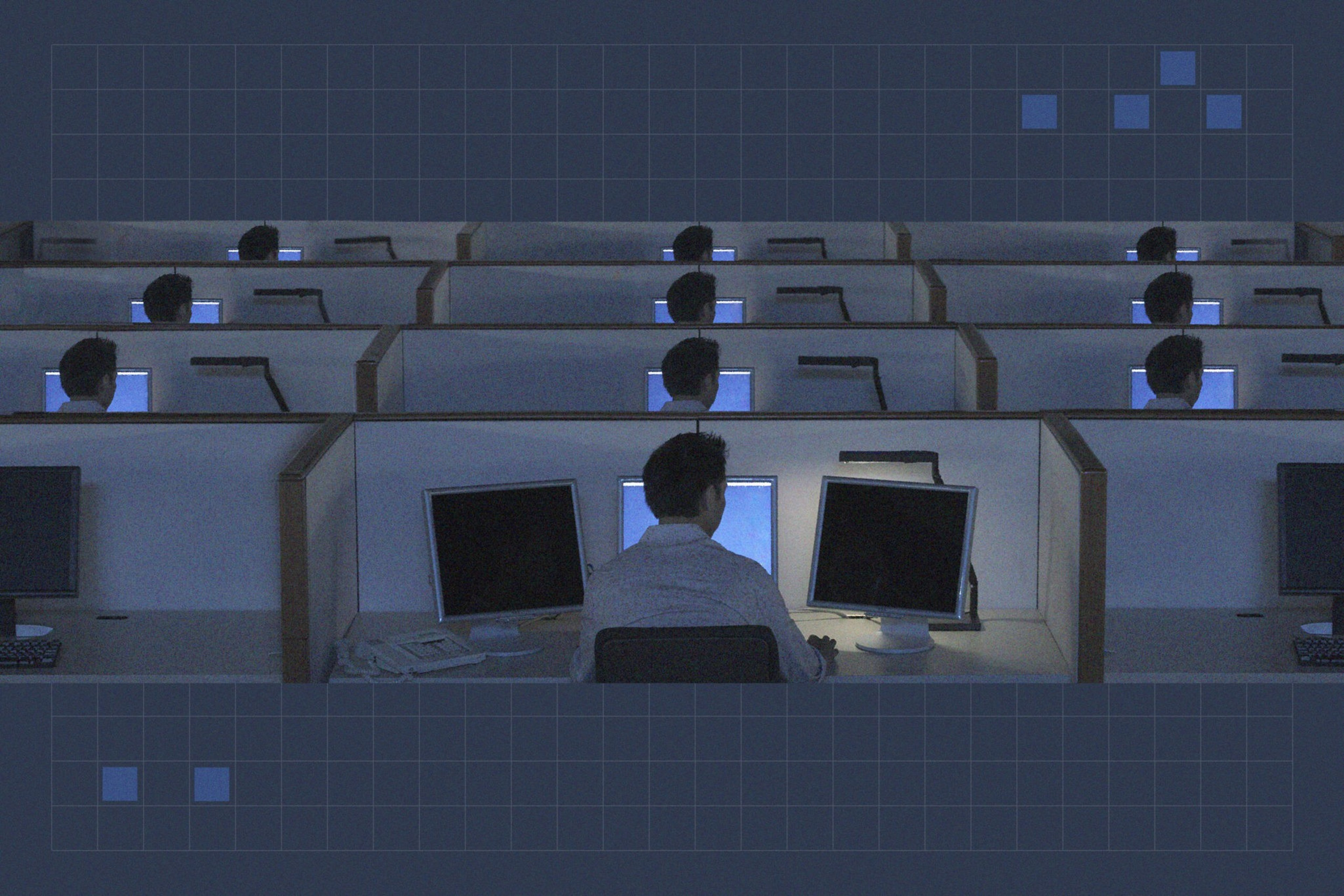 A dimly lit office with multiple cubicles, each occupied by a person facing a computer screen, arranged in rows.