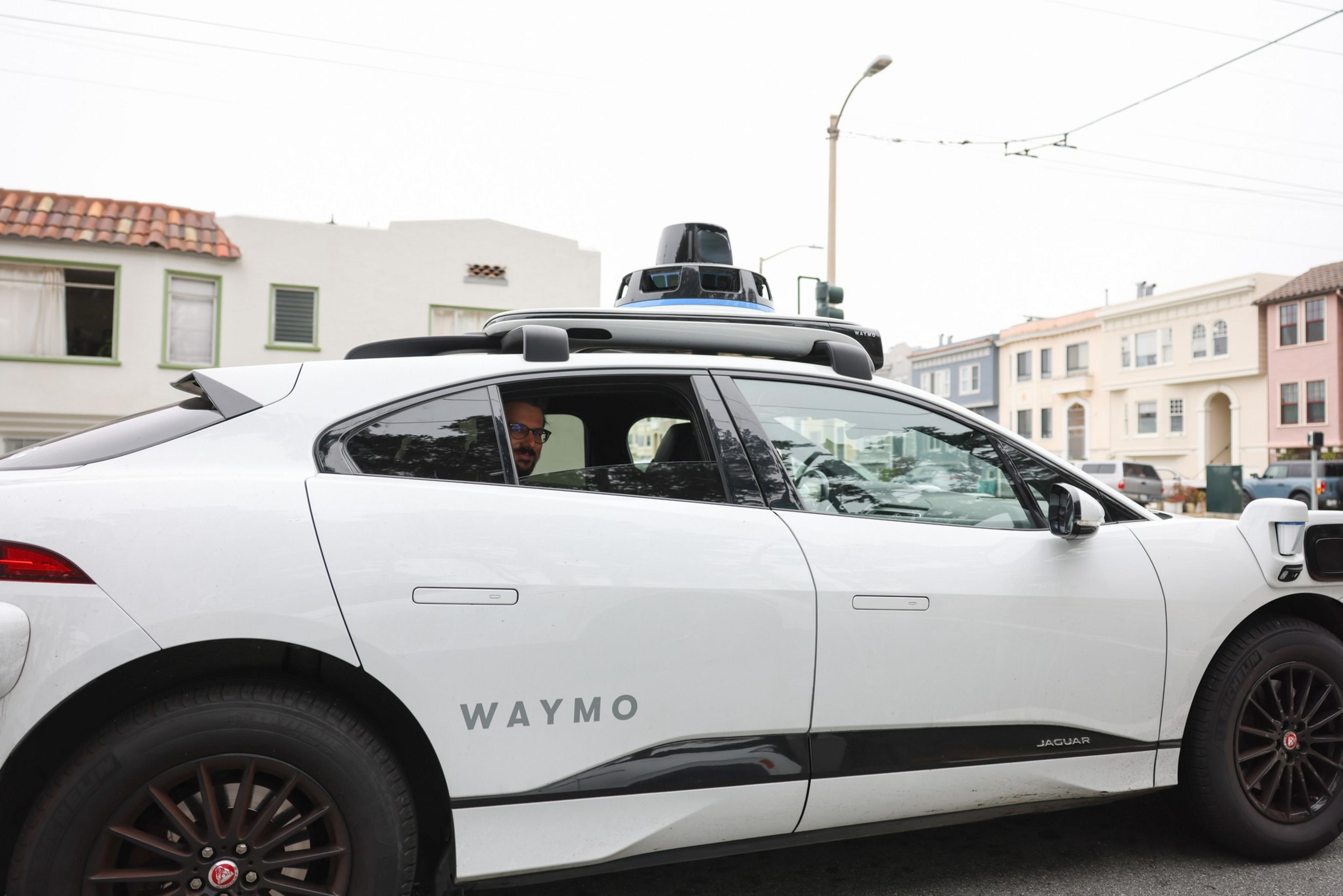 A white Waymo self-driving Jaguar car is parked on a street with a man visible inside the driver's seat.