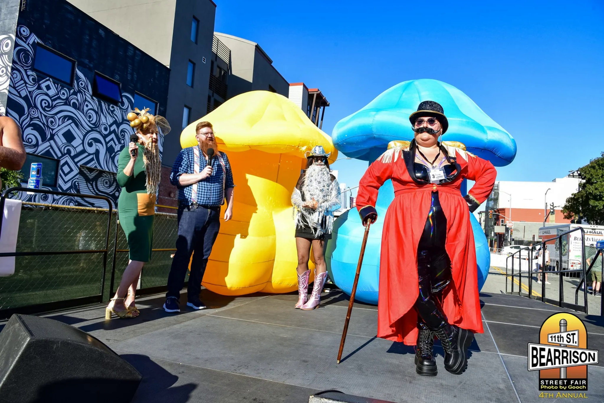 Four people stand on a stage with large yellow and blue mushroom props; one person in a red coat and top hat poses with a cane.