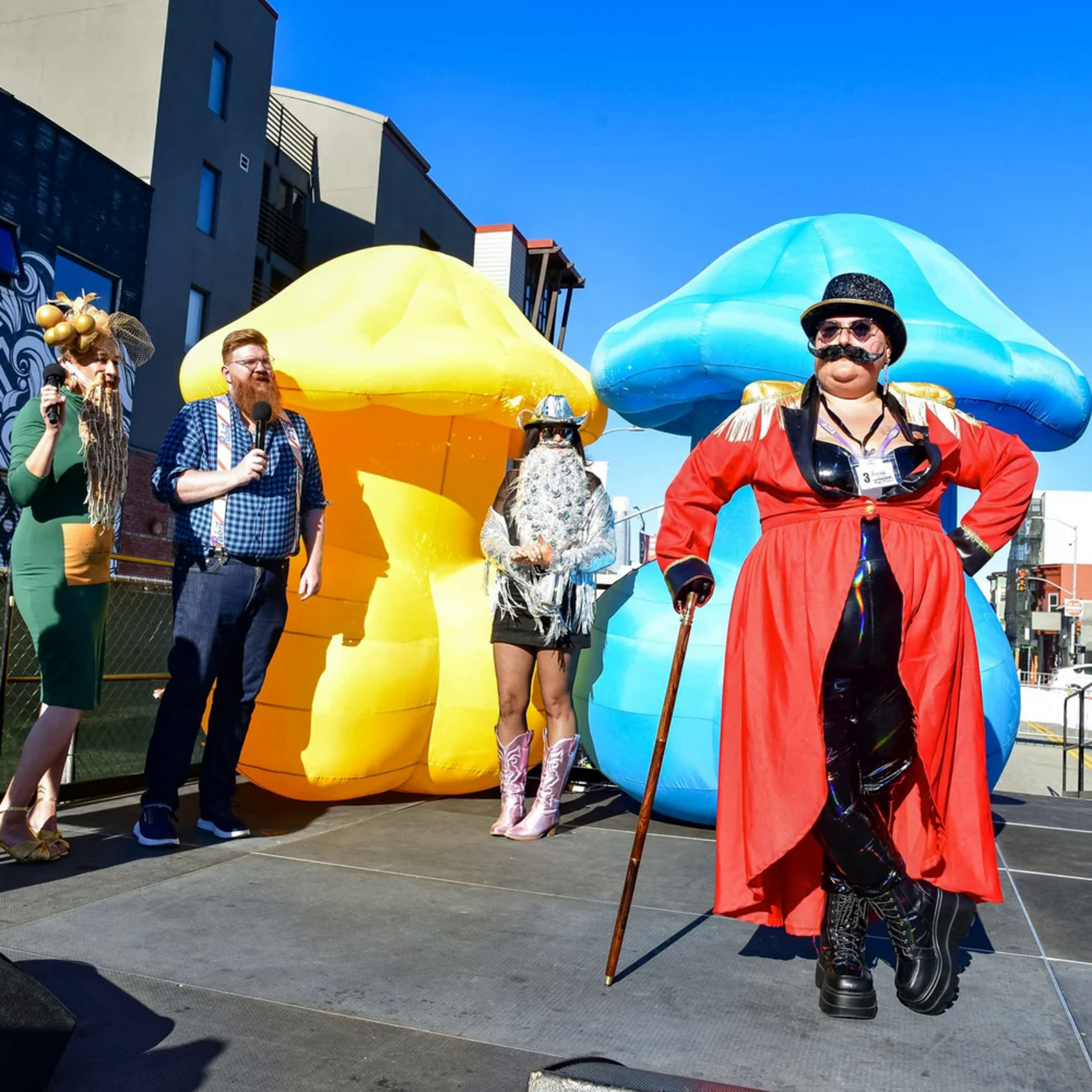 Four people stand on a stage with large yellow and blue mushroom props; one person in a red coat and top hat poses with a cane.