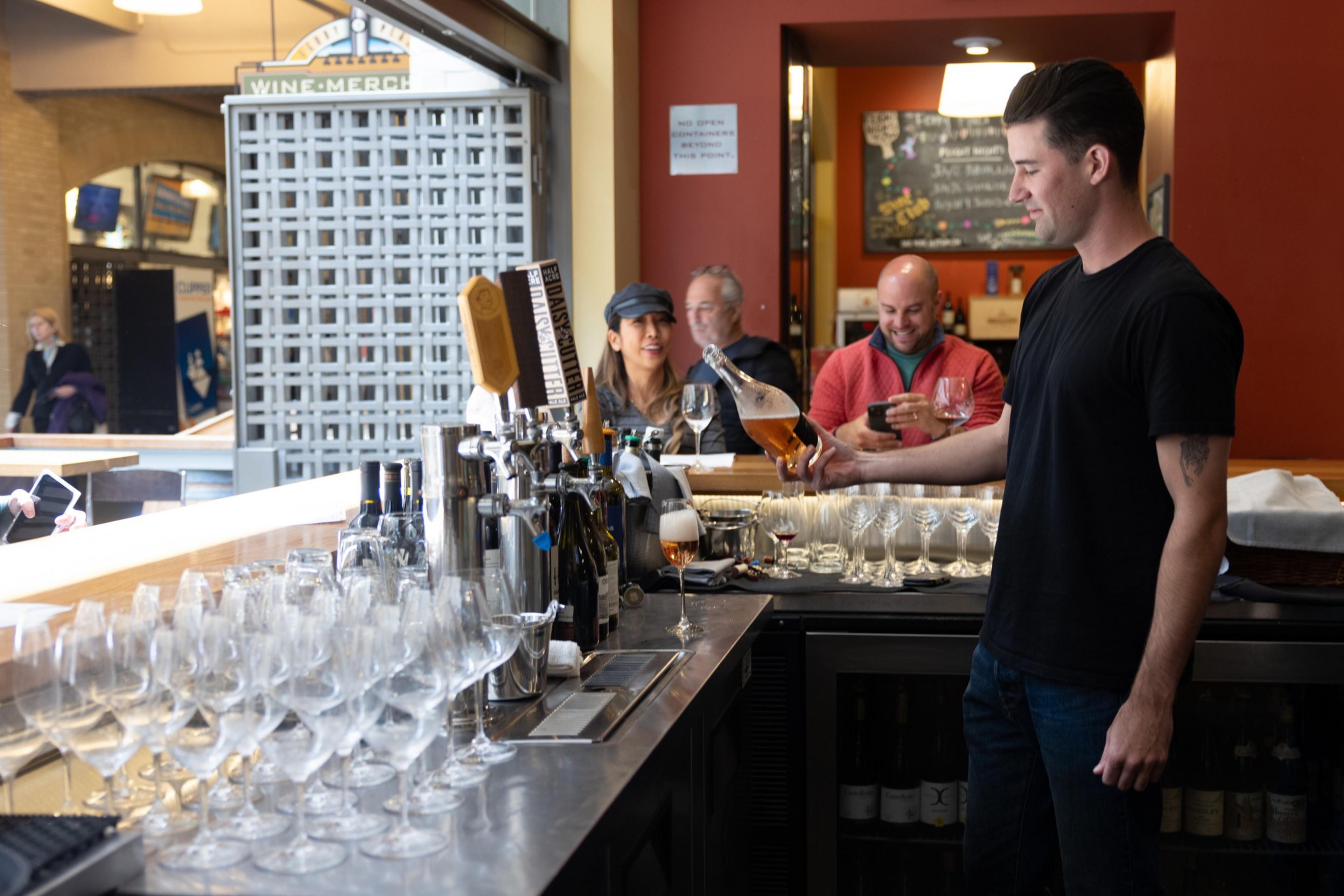 A bartender in a black shirt pours a drink behind a bar lined with clean glasses, while three patrons sit and chat in the background.