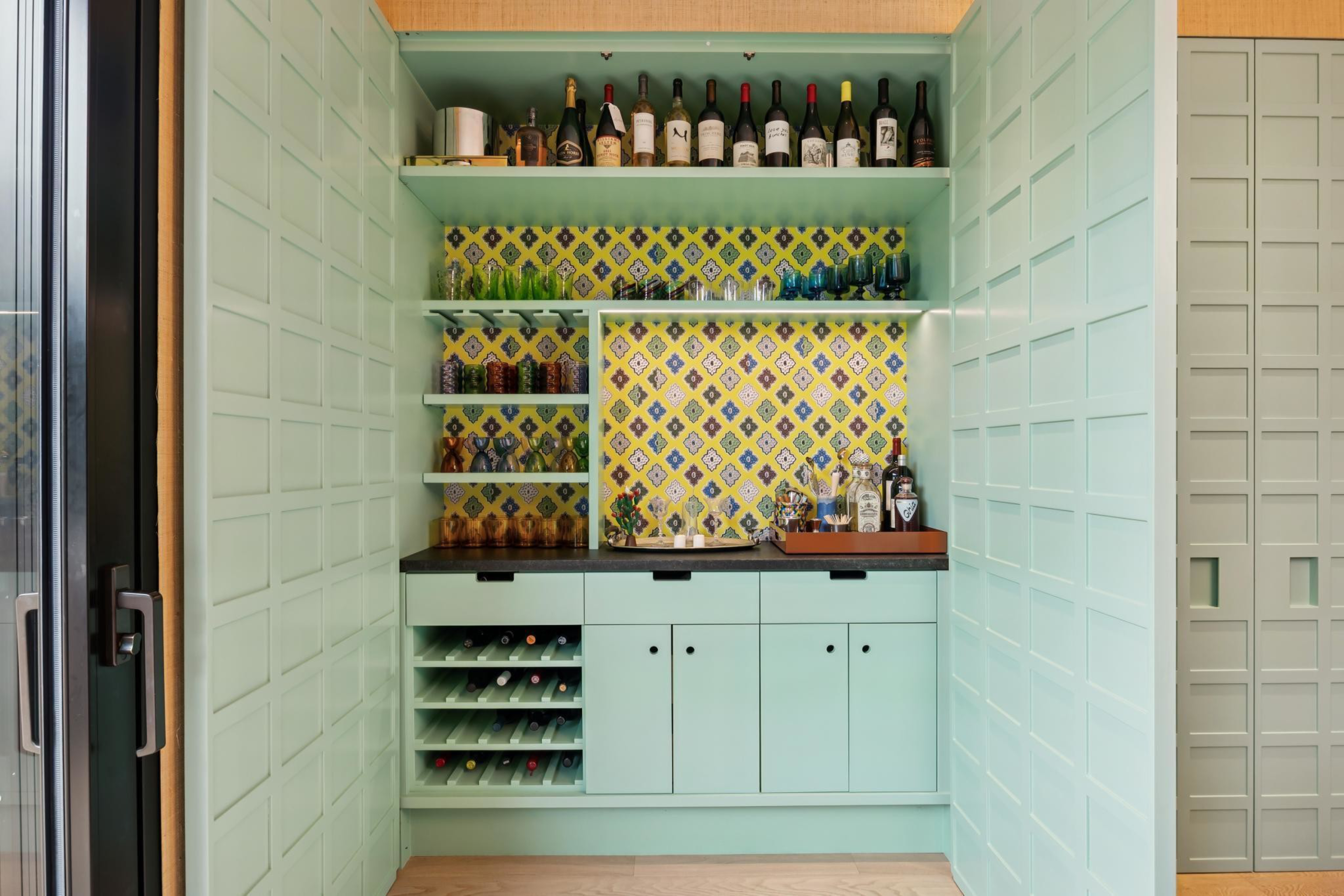 Mint green built-in bar with multiple shelves holding wine bottles, glasses, and liquor, set against yellow and blue patterned backsplash.