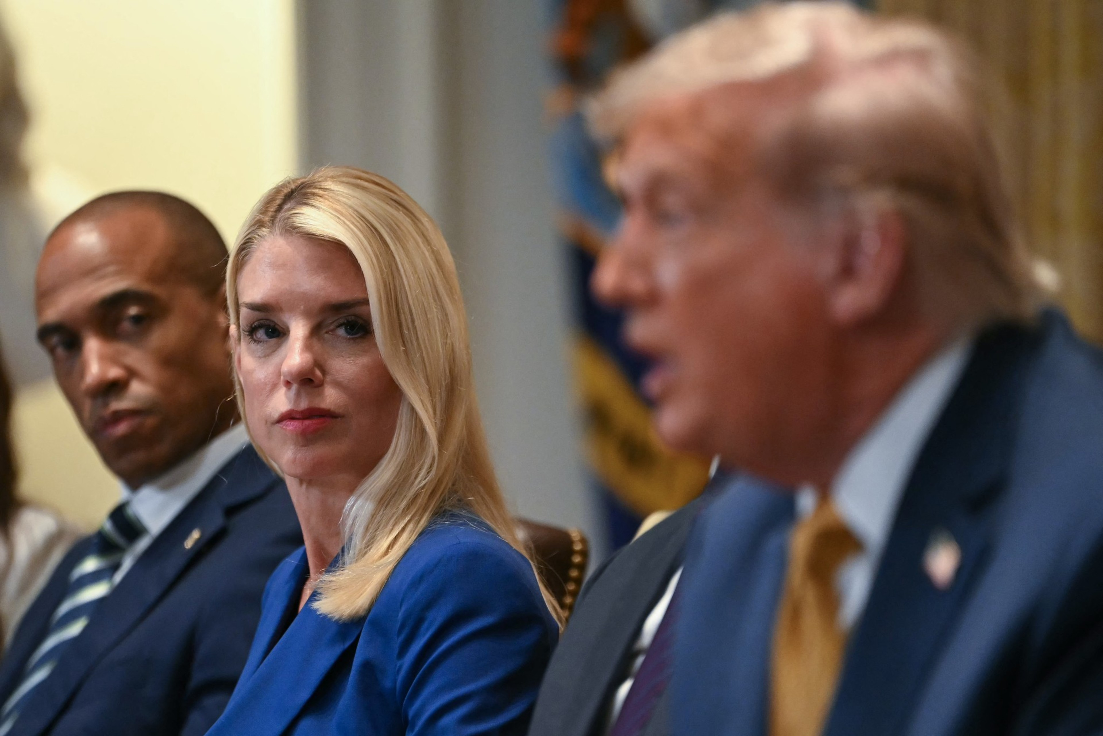 A woman in a blue suit sits between two men in suits. She looks intently at one of them, who is blurred and speaking. The background features a flag.