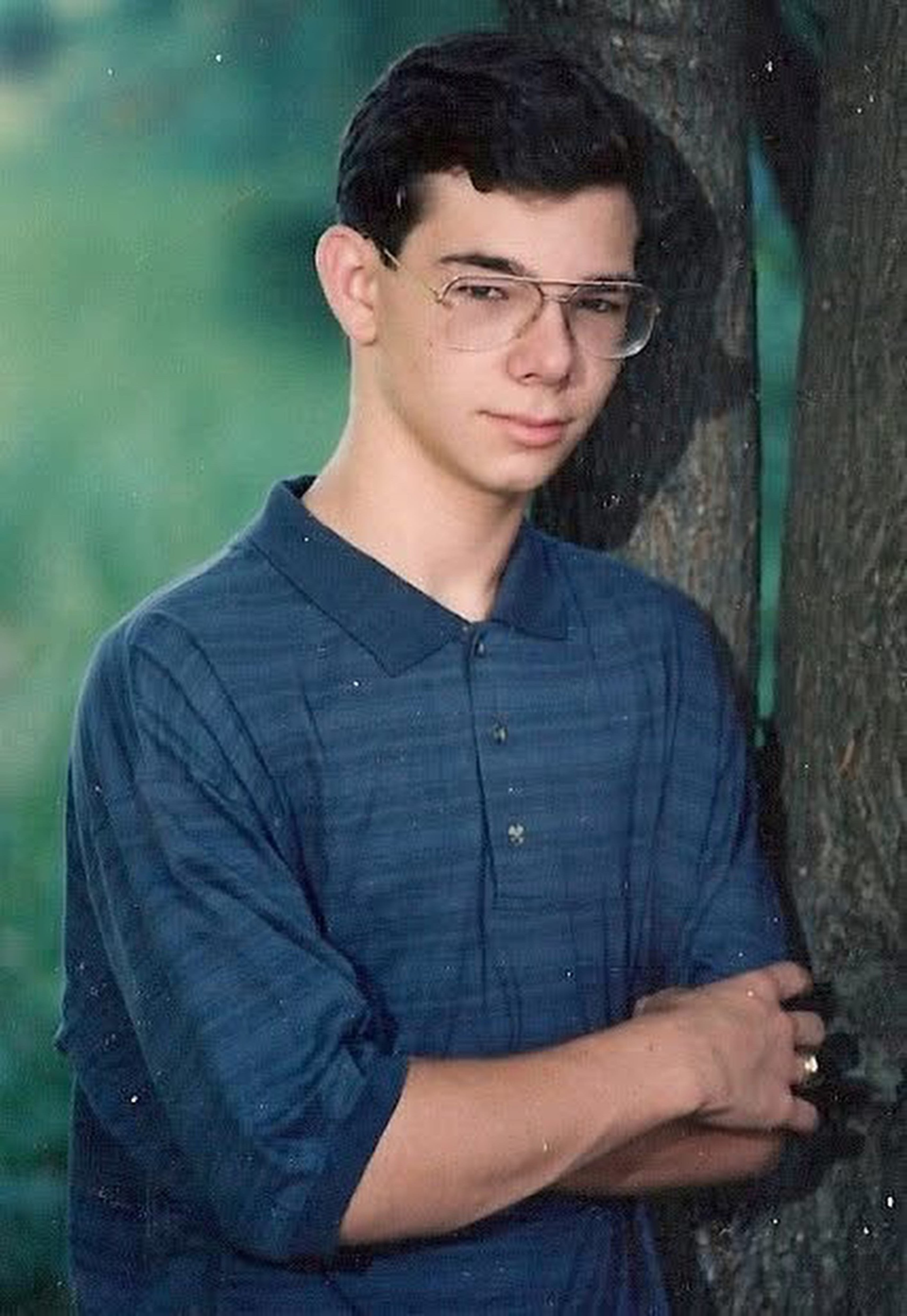 A young man with dark hair and glasses leans against a tree, wearing a blue collared shirt with his arms crossed, looking at the camera with a slight smile.